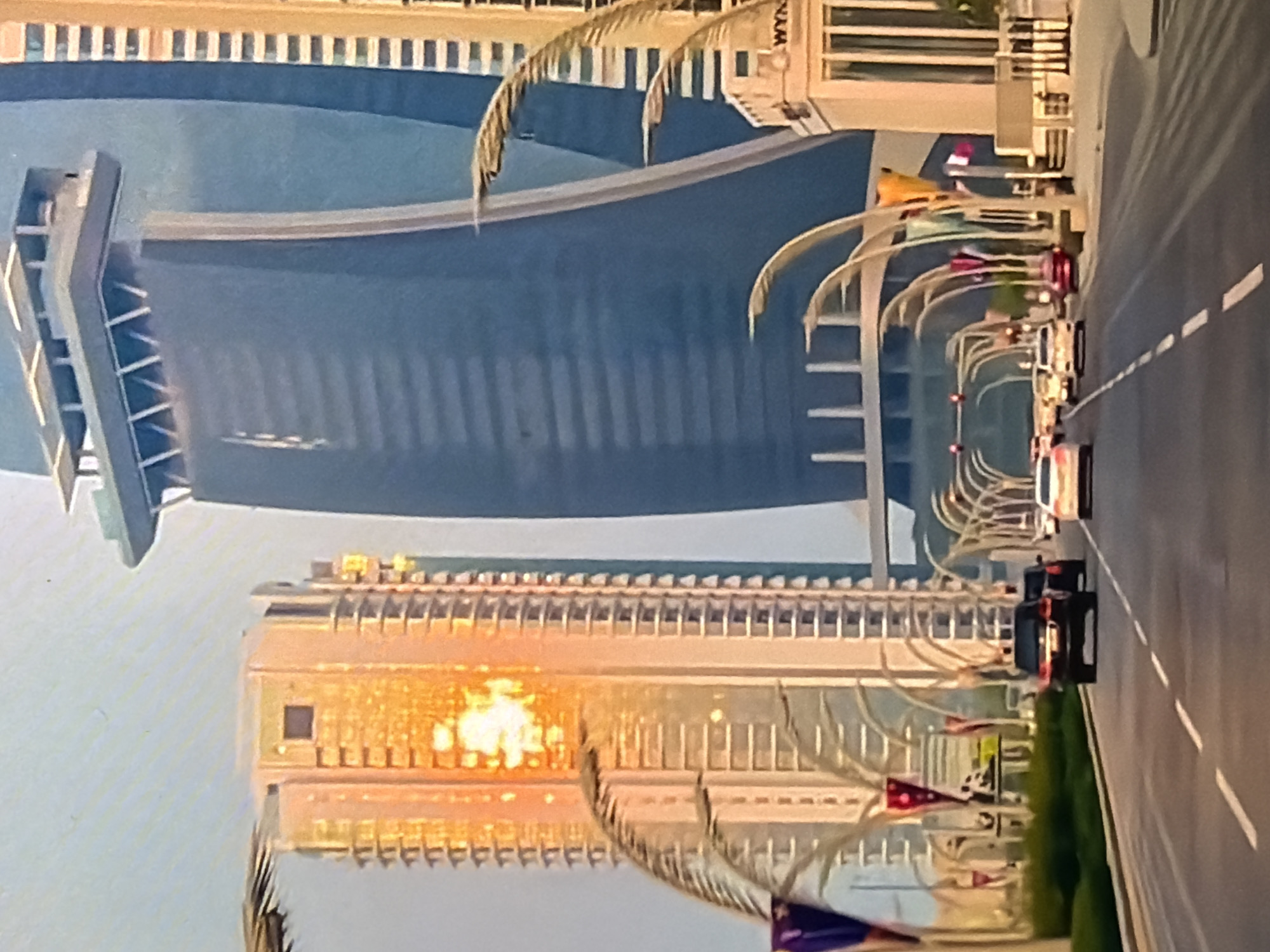 The image depicts a sunlit city street with modern architecture and vehicles. A tall, dark blue skyscraper dominates the background, characterized by its curved facade and a distinctive multi-layered structure at its apex. To its left, a narrower, lighter-colored skyscraper is illuminated by the sun, reflecting a bright orange glow. The street is paved and has multiple lanes, with several cars driving on it. Some of these cars are black SUVs, while others are lighter-colored sedans. The foreground shows a grassy median with decorative lampposts that resemble stylized palm fronds, some adorned with flags. The sky is a pale blue. The scene conveys an urban environment, likely a business or commercial district. The location of Bukuru, Nigeria is not discernable from the visual information provided in the image.