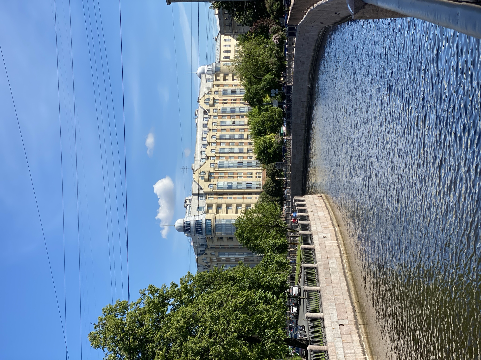 The image, oriented correctly, depicts a sunlit urban scene along a canal in Saint Petersburg, Russia, on a clear day. The canal dominates the right side of the frame, its surface a mosaic of sparkling blue and dark ripples reflecting the sky. A grey stone embankment with an ornate dark railing runs alongside the canal, receding into the distance. Several small, distant human figures can be seen on the embankment, suggesting people are strolling or cycling.
On the left side of the canal, beyond the embankment, a row of lush green trees partially obscures a grand, light-colored building with multiple stories. This building features a distinctive, curved facade, numerous windows, and a prominent domed structure topped with a slender spire on its roof. The architectural style appears classical or early 20th-century. Cars are parked along the street between the trees and the building, though they are mostly hidden by foliage.
The upper left portion of the image is filled with a bright, clear blue sky, interrupted by several thin, dark power lines stretching across the frame. A few scattered white, fluffy clouds are visible against the blue. The strong sunlight casts reflections on the water and highlights the architectural details of the building, creating a pleasant and bright atmosphere. No discernible text is visible in the image.