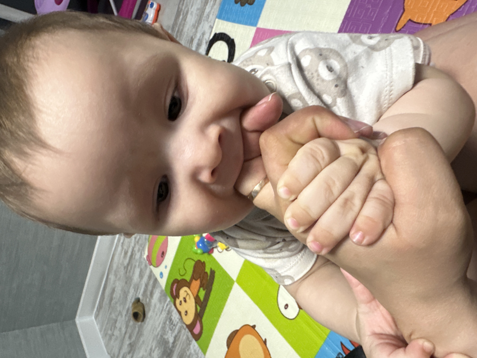An interior photograph taken from a high angle captures an infant or toddler and an adult's hands. The infant, positioned centrally, has light hair and dark eyes, and is wearing a light-colored long-sleeved top patterned with gray teddy bears. The infant is actively sucking on the ring finger of an adult's hand, which is adorned with a plain gold-colored band. The infant's small hands are holding onto the adult's hands. The adult's hands have short, unpolished fingernails. The subjects are situated on a multi-colored foam playmat featuring cartoon animal designs, including a brown monkey on a green square and a pink hippopotamus on a pink and green square. The playmat rests on a light wood-patterned floor. In the upper left background, a light gray textured wall with a white baseboard is visible. A small, dark brown cylindrical object is on the wooden floor in the upper left. A colorful beaded toy is partially visible on the playmat to the left of the infant. The setting is an indoor residential environment in Unknown, Unknown.