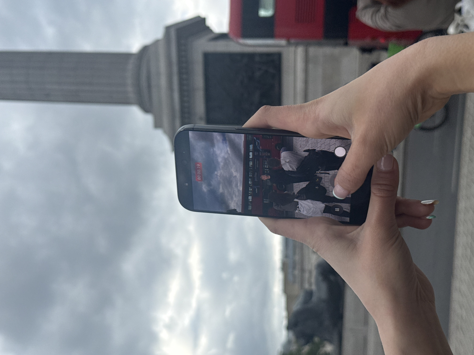 A pair of hands holds a smartphone in front of Nelson's Column in Trafalgar Square, London, United Kingdom. The phone screen shows a recording of a red double-decker bus, and people standing nearby. The column is partially visible in the background along with overcast sky.