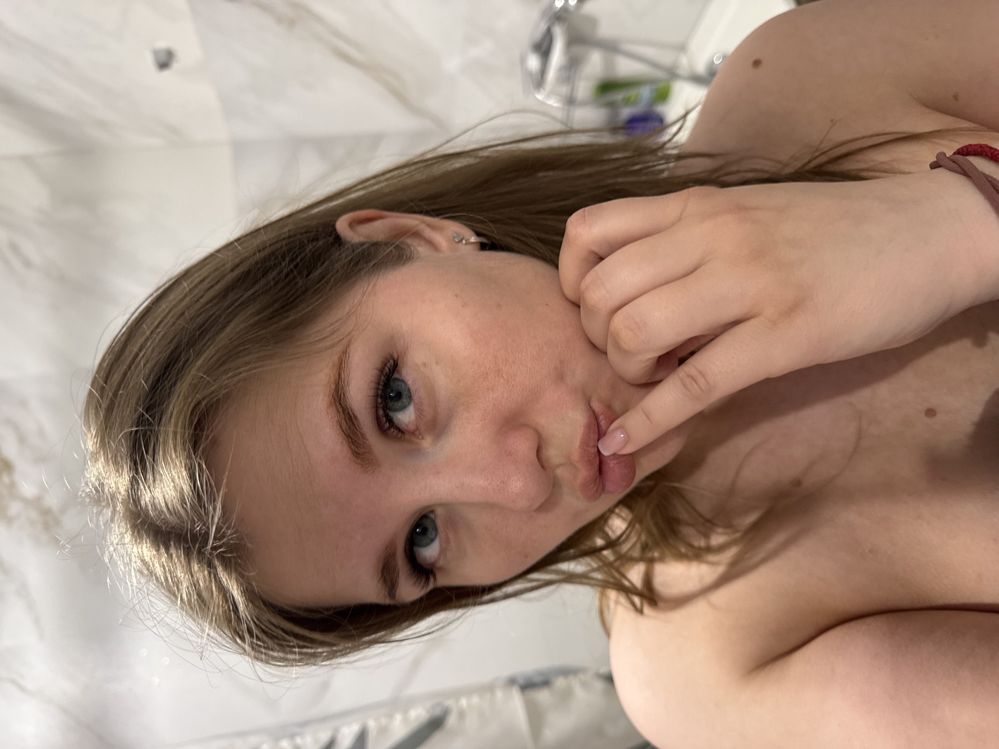 A young woman with fair skin, light brown hair, and blue eyes is posing for a selfie in what appears to be a bathroom. She is holding her finger to her lips in a playful gesture. The background shows a marble-like wall and a blurred shower fixture. It's impossible to determine the location in the city of Ферма, Russia, based on this image alone.