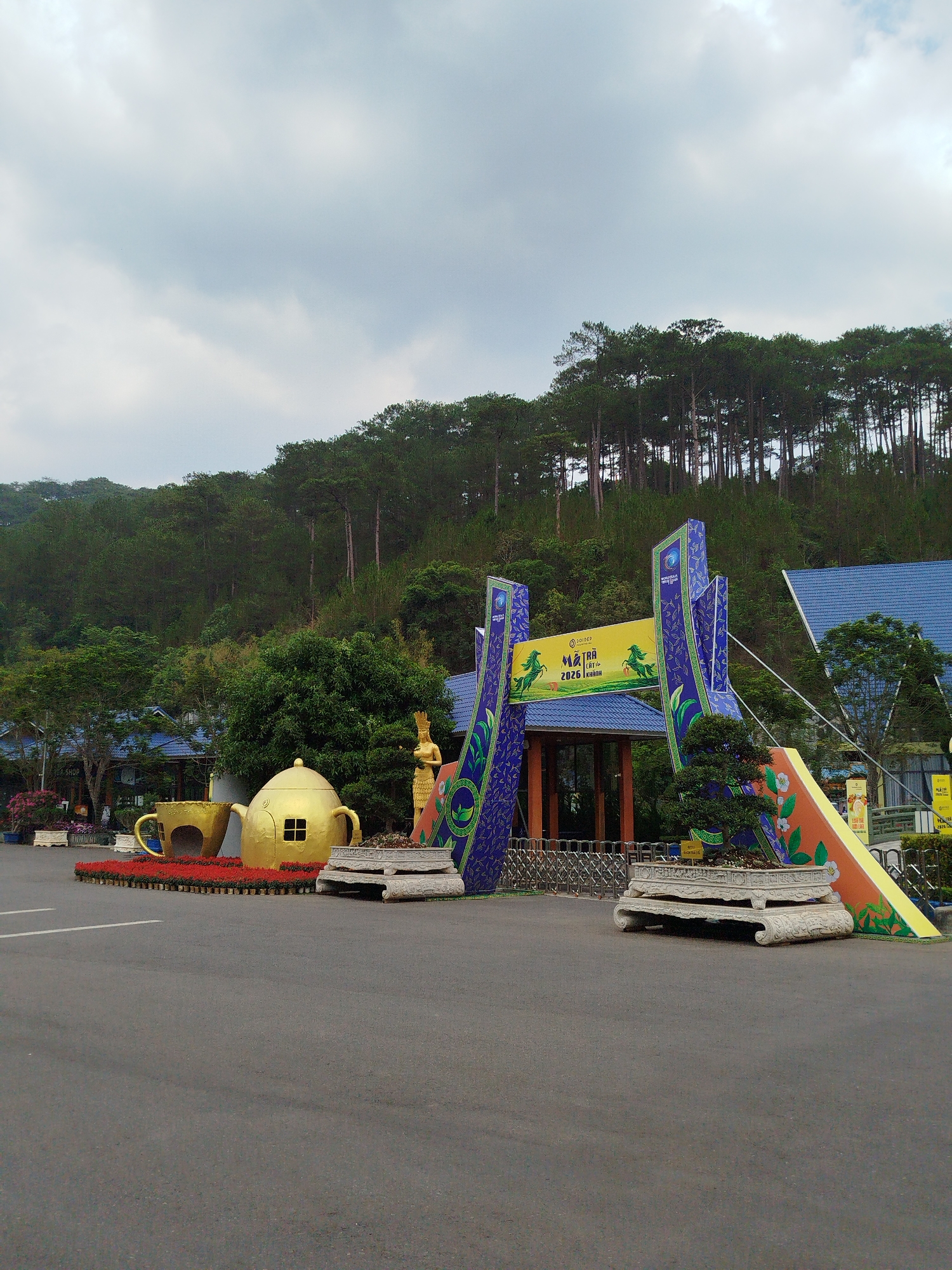 The image depicts an entrance area at an unspecified location in Hiep Thanh, Vietnam. A large, ornate archway constructed of blue and yellow material stands at the entrance. To the left of the archway, there are oversized decorative props of a gold teapot and a gold teacup placed on a bed of red flowers. In the background, a dense evergreen forest rises on a hillside. The sky is overcast with grey clouds. The entrance area is paved with asphalt, which shows signs of wear. There are no people visible in the image. The architectural style of the surrounding buildings features blue roofs. A small sign or marker is visible on the ground to the right of the archway. The overall scene suggests a tourist attraction or theme park.