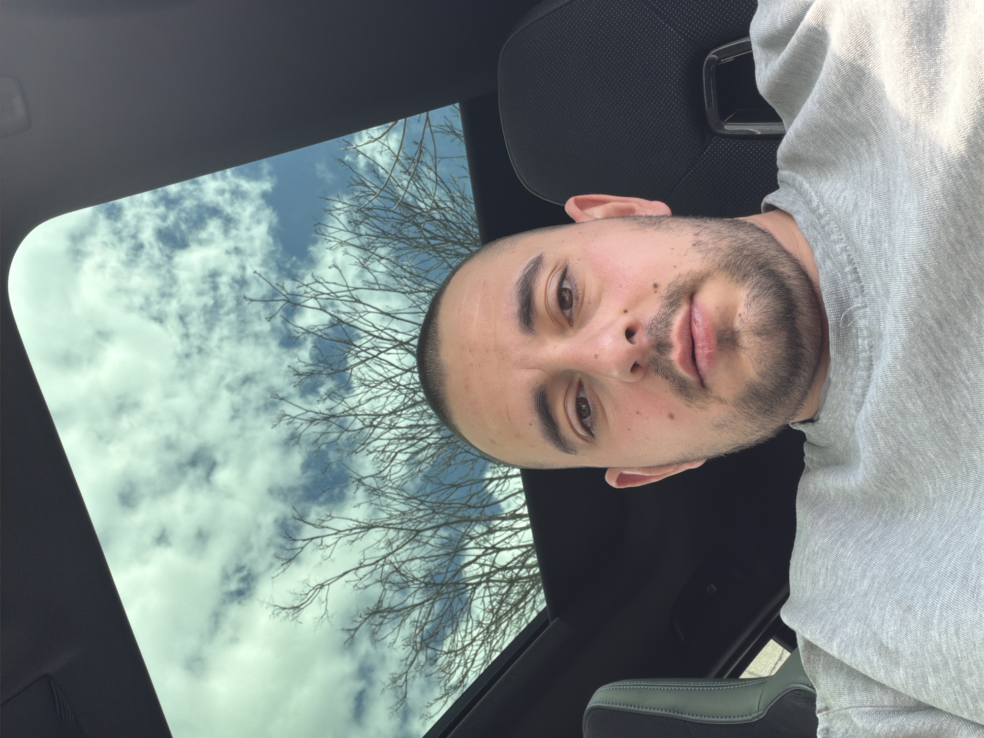 The photograph depicts a man in a vehicle, likely a car, given the presence of a black headrest and part of the dashboard. The man has a shaved head, brown eyes, and a light beard. He is wearing a grey sweatshirt. Behind him, visible through the car's sunroof or window, are bare tree branches against a sky with scattered clouds. The location is Unknown, Unknown.