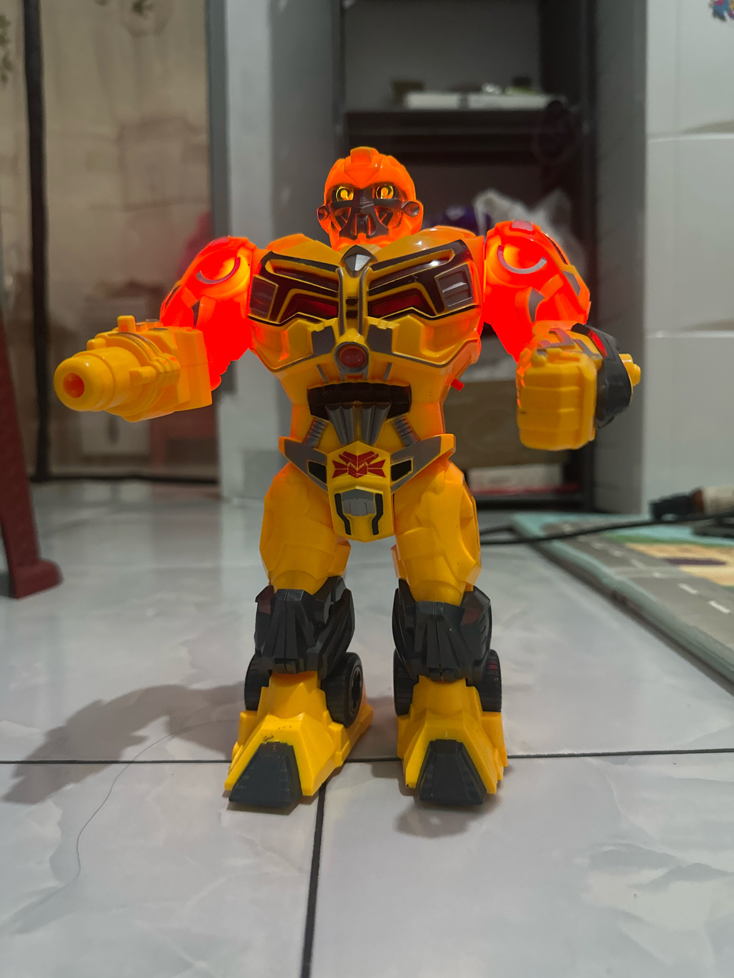 A yellow and orange robot toy is standing on a tiled floor. The robot has glowing orange parts on its shoulders and chest. It has a gun-like appendage on its left arm and a clenched fist on its right arm. The background is blurred and shows shelves and what appear to be household items. The scene is indoors, and based on the location provided, it is in a residential area of the City of Binjai, Indonesia.