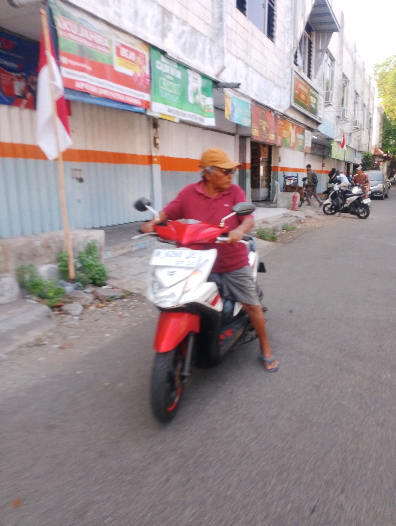 A medium shot captures a man on a red and white scooter on a street in Probolinggo, Indonesia. The man, wearing a maroon polo shirt and a tan hat, is facing left and looking ahead. He has grey hair and is wearing grey shorts and blue flip-flops. The scooter is white with red accents and has a license plate with the inscription "W AZ08 DE". In the background, a white building with orange and grey trim stands with several windows and air conditioning units visible. A red and white Indonesian flag is mounted on a pole to the left of the building. Further down the street, other people are seen on scooters and a car is parked. The street is paved and there are some green plants and debris on the side of the road. The lighting suggests it is daytime, possibly late afternoon due to the soft light.