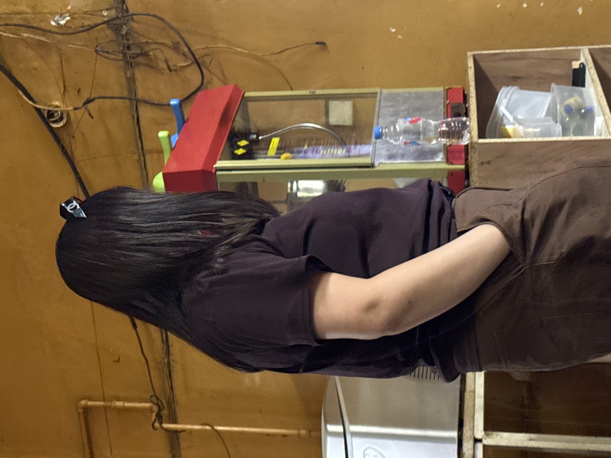 A person with long black hair stands with their back to the camera, facing a red and yellow popcorn machine. They are wearing a dark purple t-shirt and brown pants, with one hand in their pocket. The popcorn machine has a clear glass front, showing internal workings, and a bottle of water sits on top. Behind the person, a yellow wall is visible, with pipes and electrical wires running along it. In the foreground, a wooden shelf unit holds two clear plastic containers. To the left of the popcorn machine is a white appliance, possibly a refrigerator. The scene appears to be indoors, with artificial lighting.