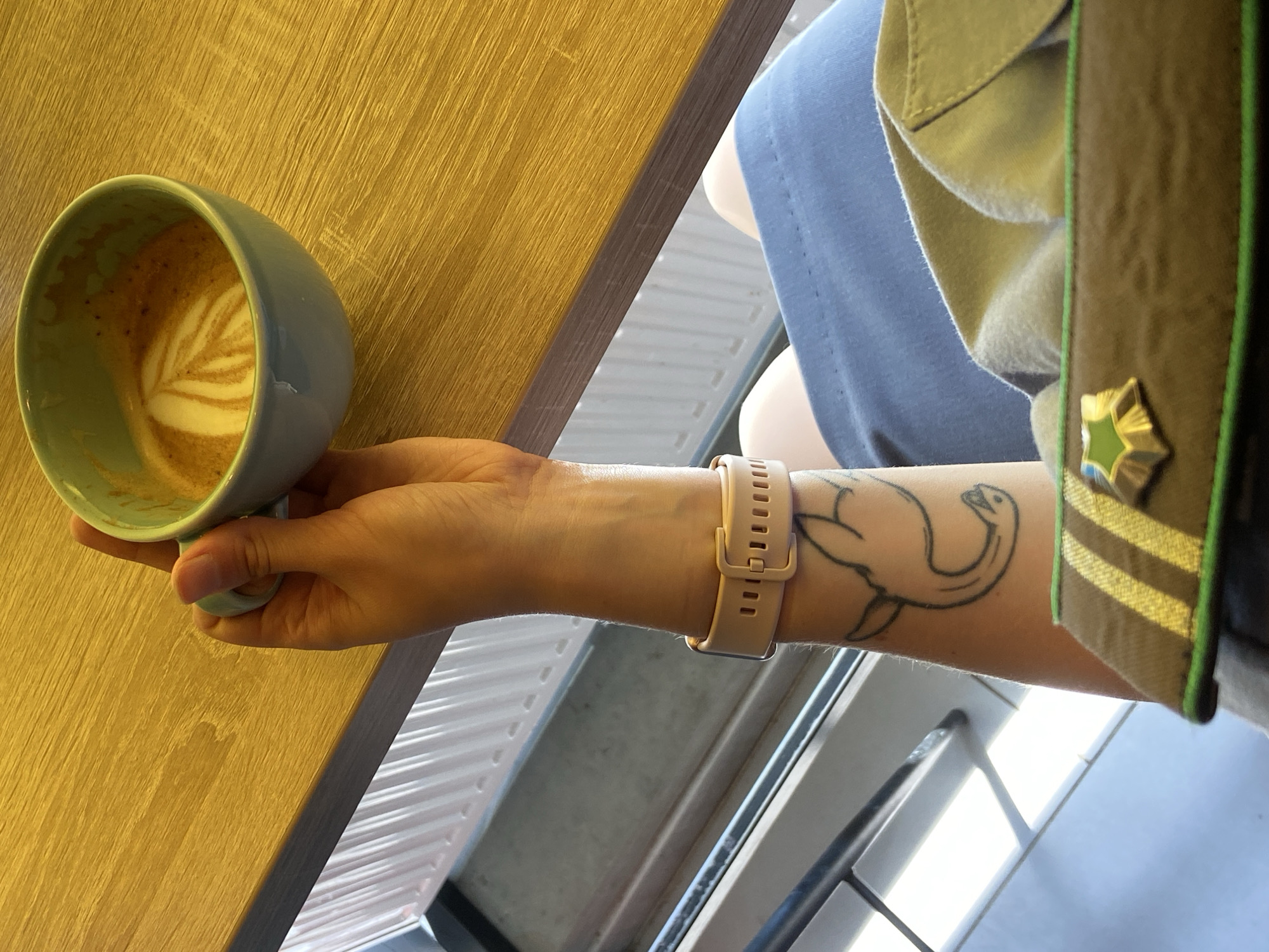 A person's left arm, adorned with a tattoo of a stylized animal and a light pink smartwatch, is visible. The arm holds a light blue mug filled with coffee and latte art. The mug rests on a light brown wooden table. In the foreground, a portion of a khaki-colored garment with green and yellow stripes and a star-shaped emblem is visible. A blue skirt is also visible on the person. The background shows a white radiator and a window. The scene is indoors, and no specific location within Tyumen, Russia, can be determined.