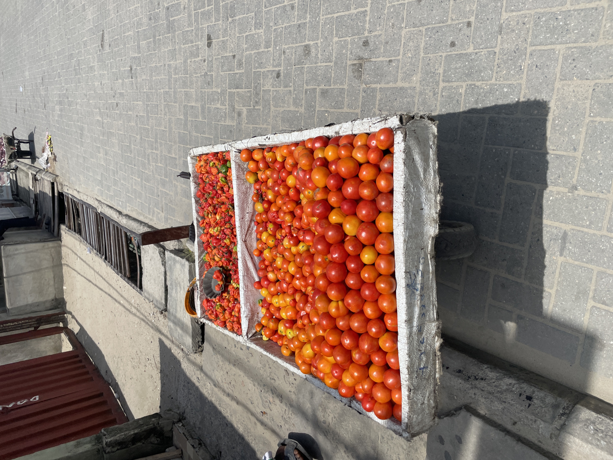 An elevated, high-angle shot captures a roadside or market scene, primarily focusing on two large, rectangular crates overflowing with fresh produce. The scene is set on a paved surface made of interlocking grey bricks, with a strong, distinct shadow cast by one of the crates indicating bright sunlight, likely mid-morning or afternoon.
The crate on the right is filled with a dense array of ripe, round tomatoes, predominantly in shades of red and orange, with some yellower ones mixed in. The crate on the left holds a large quantity of smaller, vibrantly colored peppers, mostly red and orange with hints of green, resembling habanero or scotch bonnet peppers. A small, grey metallic bowl or scoop is placed within this crate of peppers.
Below the level of the crates, to the left, are concrete structures and a metal railing, suggesting a multi-level building or an elevated walkway overlooking lower ground. A red corrugated metal roof is visible in the lower-left background. A person's foot, wearing a grey sandal, is partially visible at the bottom edge of the frame, near the crates, hinting at a vendor or customer.
Visible text is limited and partially obscured. On the side of the crate holding tomatoes, some faded blue writing, possibly "MAC KEC" or similar, can be faintly seen. On the red corrugated metal roof below, very faint letters like "DGA" are barely discernible. The overall scene depicts an outdoor produce display in Ado, Nigeria.