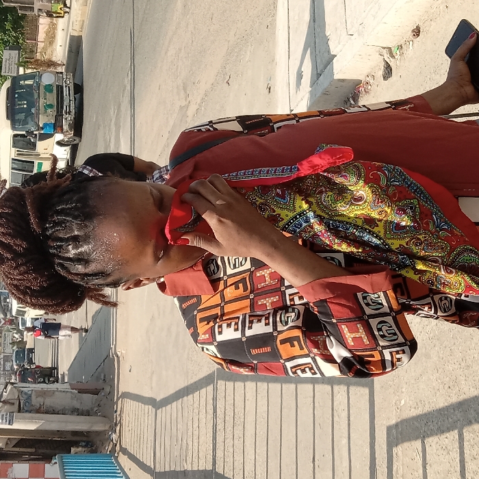 A woman with dreadlocks pulled into a bun stands on a sidewalk. She wears a colorful, patterned shirt and rust-colored pants. She holds a red cloth to her face. A colorful scarf drapes around her neck. A phone is visible in her hand. A busy street with cars and pedestrians is in the background. The scene is brightly lit with sunlight.
