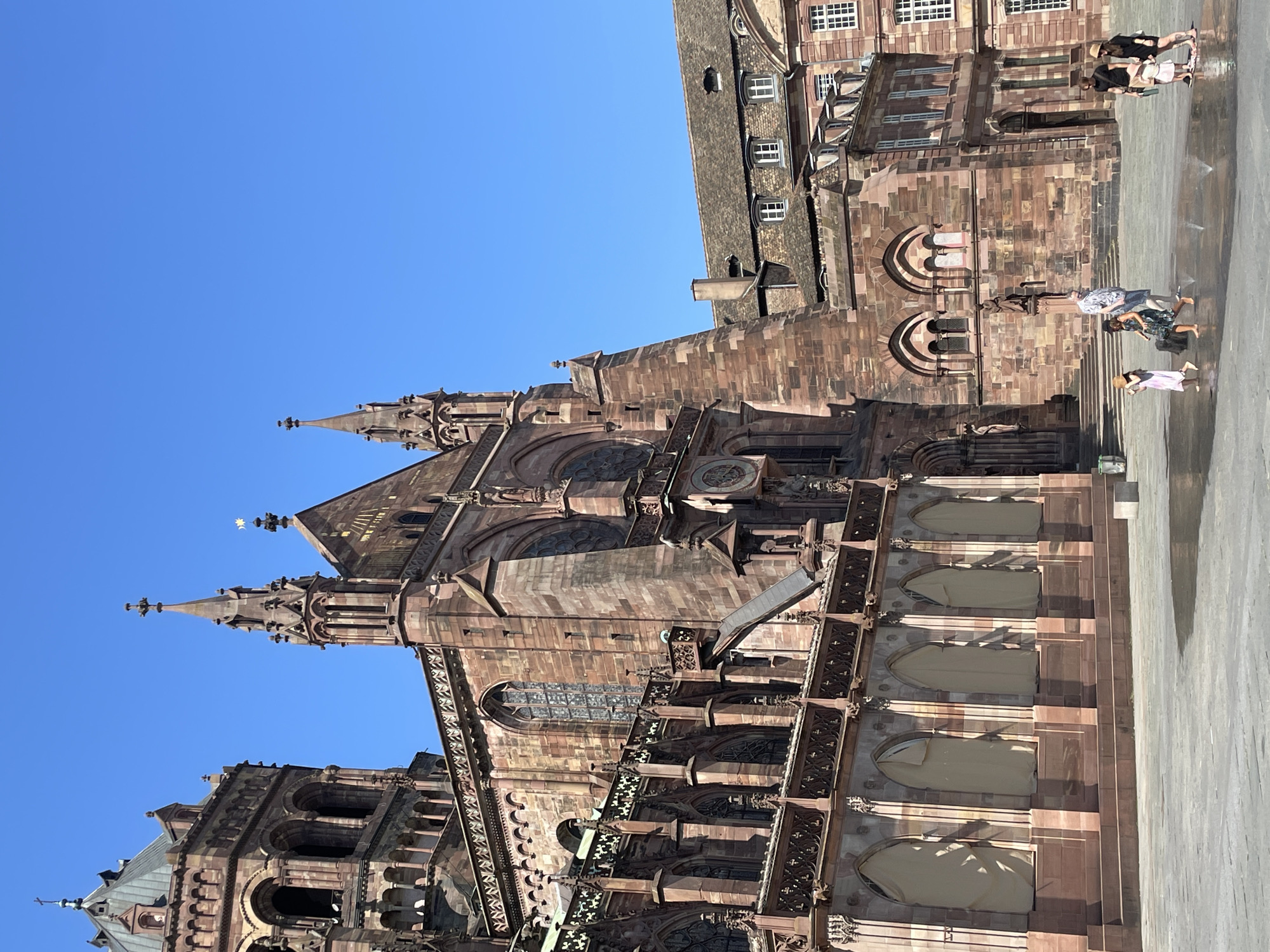 In Strasbourg, France, a large gothic cathedral constructed of red sandstone is featured prominently against a clear blue sky. The cathedral exhibits intricate architectural details, including spires, arches, and decorative stonework. In the foreground, a paved plaza is wet, indicating the presence of a water feature where several people are interacting. Two families are visible: one group consists of an adult male and female with two young children, one of whom is playing in the water. Another group comprises an adult male and female with a young child. The plaza is devoid of trees or significant vegetation.
