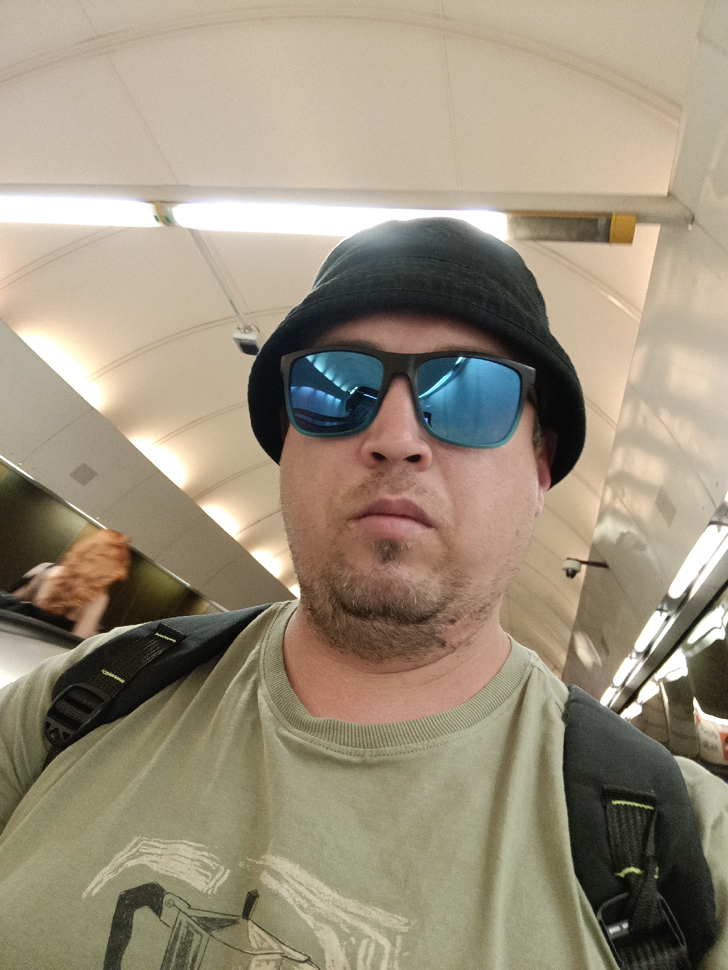 A man with light skin, a goatee, and stubble is wearing a black bucket hat and blue-tinted sunglasses. He has a light green t-shirt with a graphic print on it and a black backpack with green accents. He is looking directly at the camera. The background appears to be a modern indoor space with a curved, cream-colored ceiling and fluorescent lighting. The man is standing on an escalator, as indicated by the angled wall to his left, on which a blurry figure with reddish hair is visible. This is likely a public transit station in Prague, Czechia.