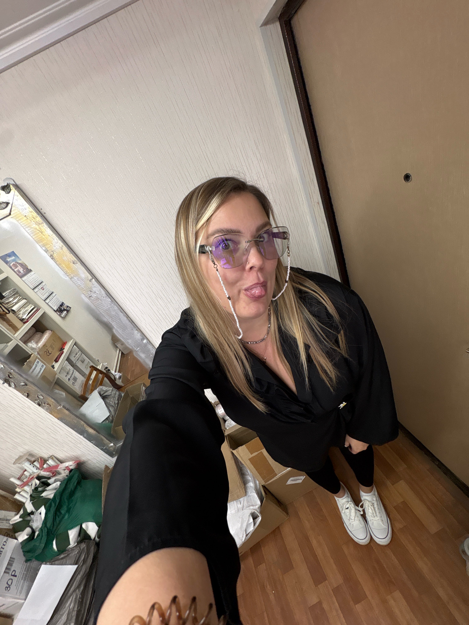 A woman, with blonde hair and wearing a black dress, black leggings, and white sneakers, is standing in a room and taking a selfie. She is wearing large, square, purple-tinted glasses with a beaded chain attached. She is sticking her tongue out at the camera. Behind her, to the left, is a large mirror reflecting a room with shelves filled with boxes and papers. To her right is a closed, light brown door with a peephole. The floor is made of polished wood planks. The image appears to have been taken indoors, possibly in an apartment in Noviy, Russia, given the context.