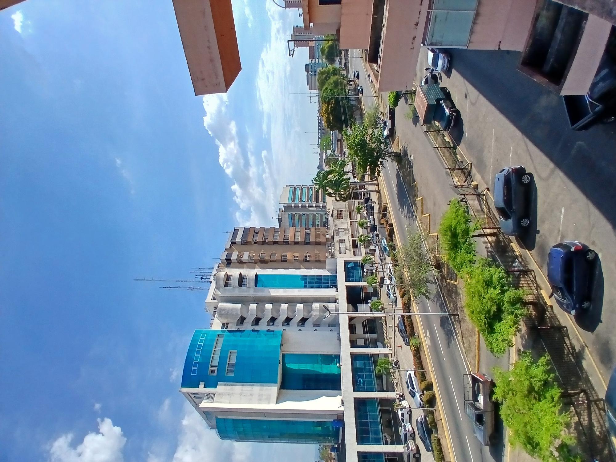 The image depicts an aerial view of a street scene in Ciudad Guayana, Venezuela. A multi-story building with a blue glass facade is visible on the left side of the frame. A street runs parallel to the building, lined with parked cars and trees. Other buildings and distant structures are visible in the background under a blue sky with scattered clouds. There are no people visible in the image.