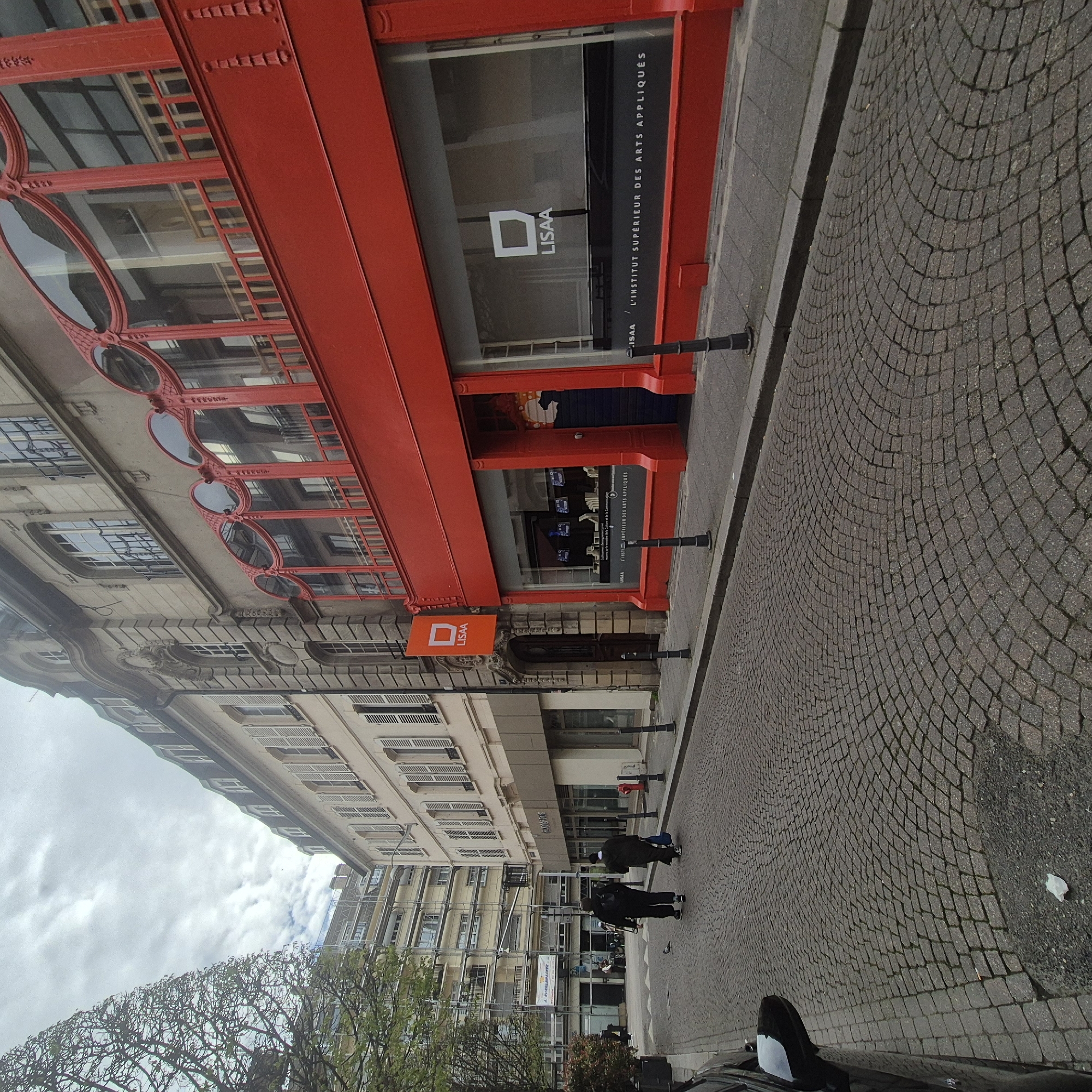 This is an outdoor shot in Strasbourg, France. The image is rotated to the left. The view is of a street scene on a cloudy day. The building to the left of the street is adorned with a red, ornately-framed structure, housing a shop. The shop's signage reads "LISAA / L'INSTITUT SUPÉRIEUR DES ARTS APPLIQUÉS." Down the street, a pair of pedestrians walk along the cobblestone path. A vehicle is seen in the lower right corner.