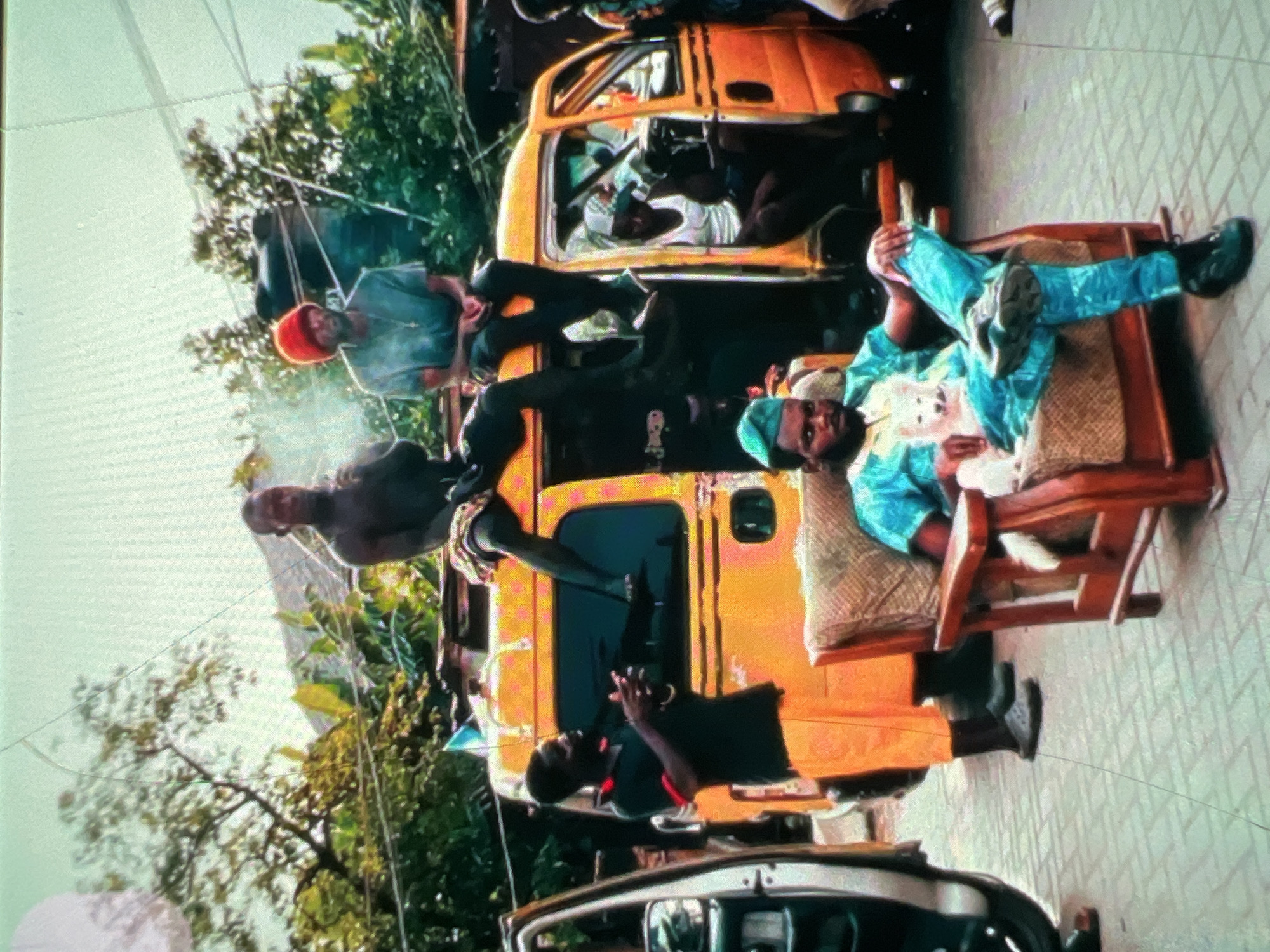 In Lagos, Nigeria, a street scene unfolds with a vibrantly colored yellow van as the central focus. The van is packed with people, some sitting on top and others inside, suggesting a mode of transport or a gathering. In the foreground, a man is seated in a wooden chair, holding a small white dog. He is dressed in a striking blue and green outfit, accessorized with a matching green cap. The surrounding environment appears to be a paved street, indicative of an urban setting. The image captures a moment of daily life, with individuals interacting and congregating around the van and the seated man, painting a vivid picture of Lagos's bustling street culture.