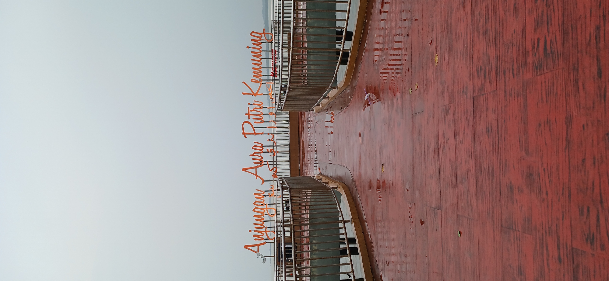 The image depicts the "Anjungan Aura Putri Kemuning" in Tanjung Balai Karimun, Indonesia. The scene is an outdoor waterfront area with a wooden boardwalk that appears wet, suggesting recent rain. The boardwalk is a deep red color with visible plank lines and some scattered leaves. Two metal railings with vertical bars line either side of the walkway, leading towards a body of water. In the background, large, orange, cursive-style lettering spells out "Anjungan Aura Putri Kemuning." The sky is overcast and a pale gray. There are no people visible in the image.