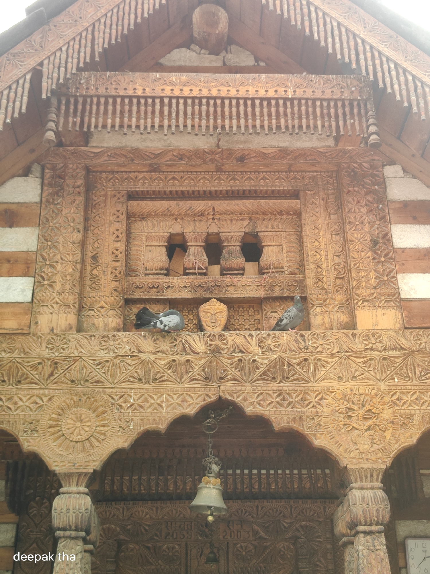 The image shows the facade of a wooden building in Vashisht, India. The building features intricate wood carvings, including decorative patterns, floral motifs, and a carved face above a central opening. Two pigeons are perched on a ledge within the opening, and another pigeon is seen to the right. A bell hangs from the ceiling of an arched entryway. The architecture suggests a traditional style common in the region. The timestamp in the bottom right corner indicates the image was taken on December 26, 2023, at 12:32 PM. A watermark in the bottom left corner reads "deepak tha". The overall scene is one of detailed craftsmanship and architectural beauty.