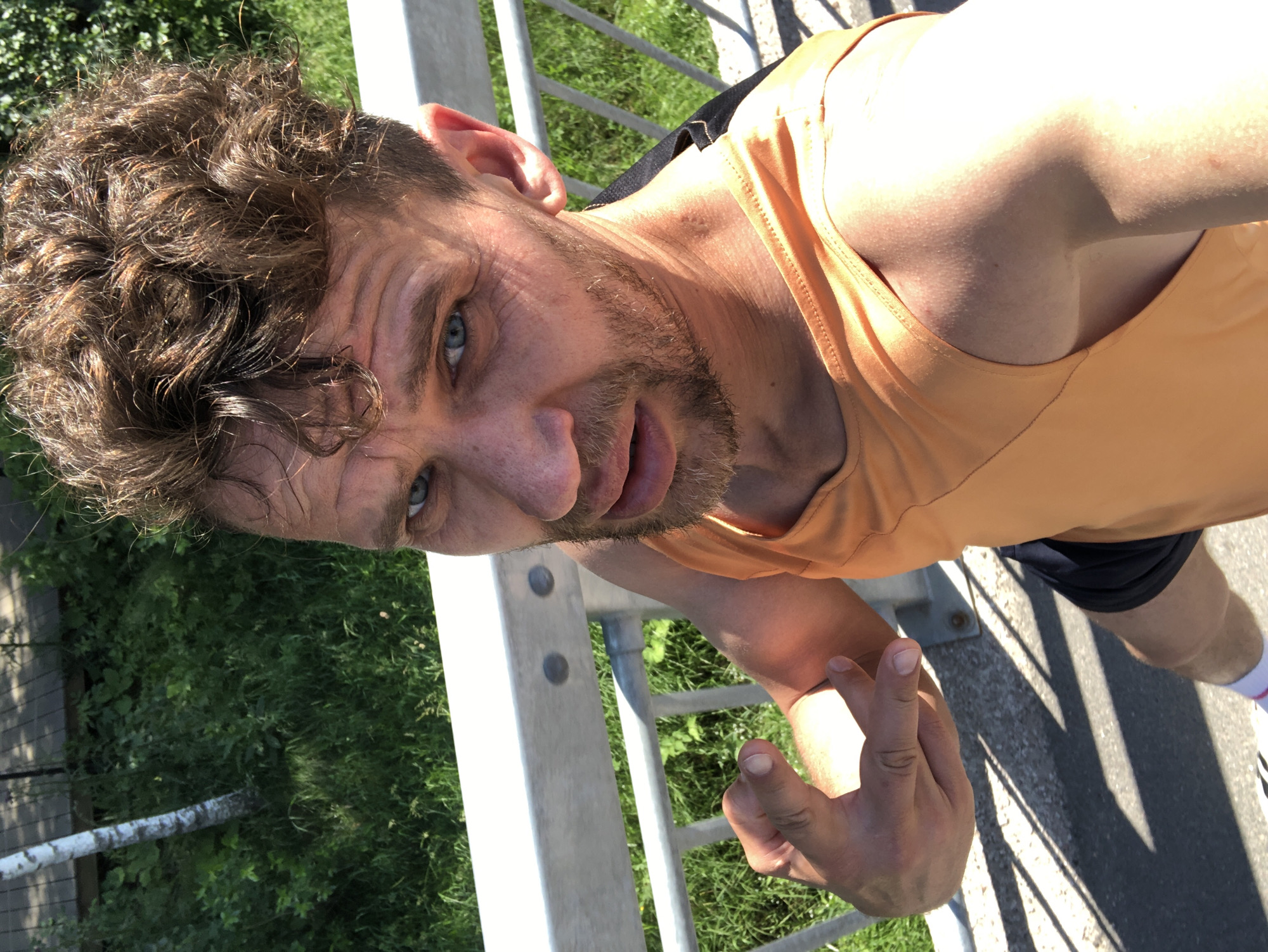 An individual is standing on a pedestrian bridge, holding up their left hand in a gesture. They have brown, curly hair and blue eyes, with stubble on their face. The individual is wearing an orange tank top and dark shorts. The background consists of greenery and a metal railing. The scene is outdoors on a sunny day. The location is identified as Saint Petersburg, Russia.