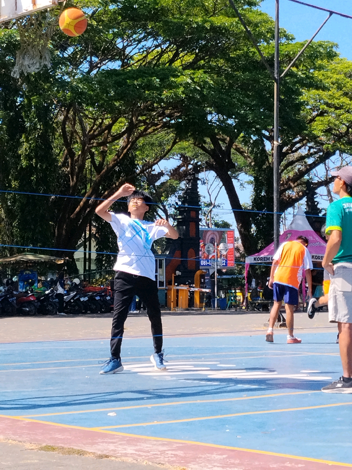 A young Asian male, wearing a white t-shirt with a blue design and black pants, is mid-action on an outdoor basketball court. He is in the process of throwing or passing a basketball, which is visible in the air above and to his left, near a basketball hoop and net. The court is blue with yellow and red boundary lines. In the background, there are several parked motorcycles, a large tree with lush green foliage providing shade, a dark-colored structure resembling a gate or entrance, and colorful tents. The sky is clear and blue. To the right, another male, wearing a teal shirt and a cap, is partially visible, as is a third person in an orange vest and blue shorts. The scene appears to be in an open area, possibly a park or recreational facility, within Malang, Indonesia, suggested by the architectural elements and vegetation.