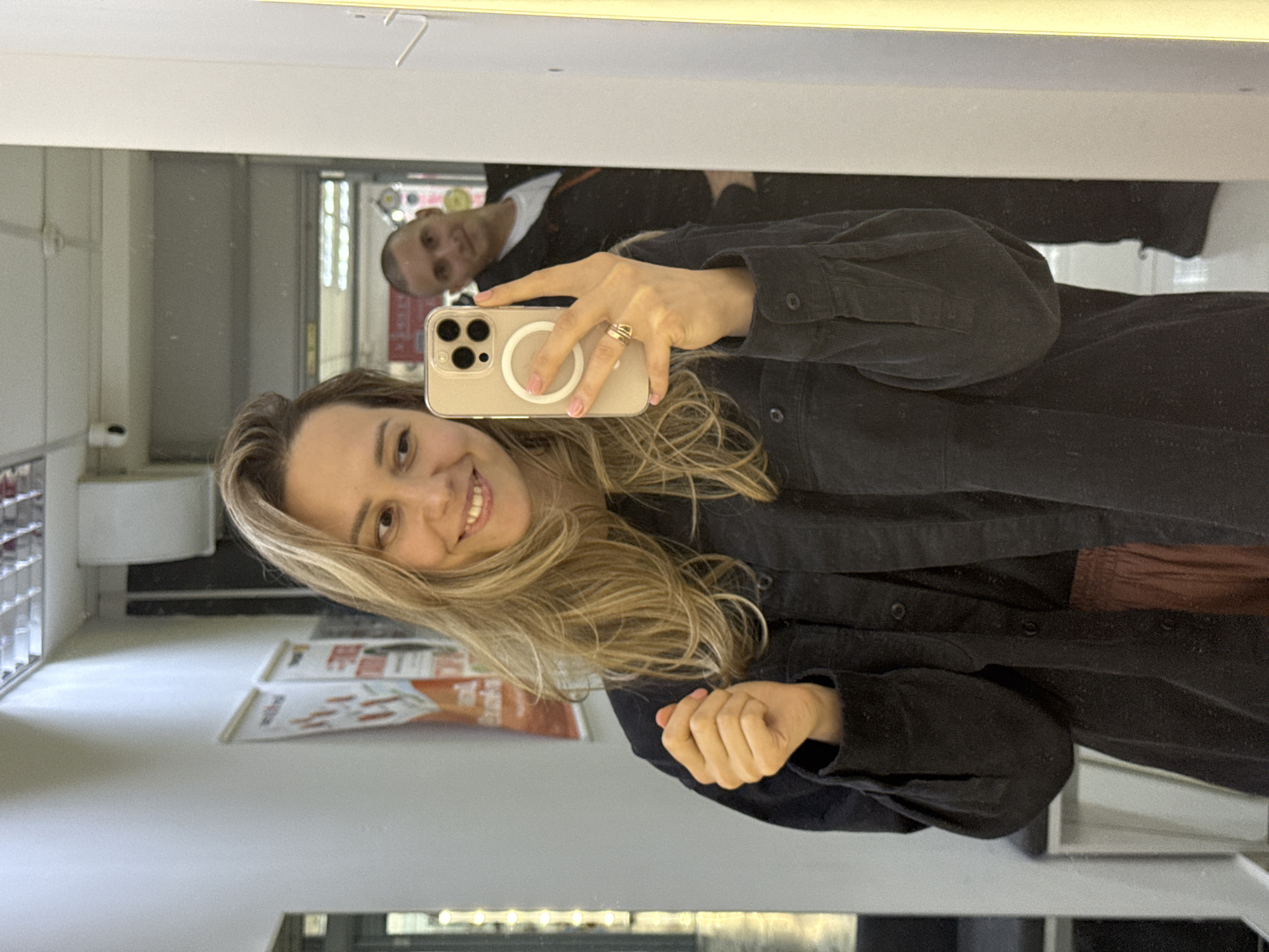 A woman is taking a mirror selfie indoors. She is smiling broadly towards her smartphone, which she holds with her right hand, capturing her reflection. She has long, wavy blonde hair and is wearing a dark, long-sleeved shirt or jacket. A gold-colored ring is visible on her right ring finger, and her fingernails feature a light pink French manicure style. The smartphone is light-colored with a prominent circular MagSafe accessory on its back.
In the background reflection, behind and slightly above the woman's head, a man with short hair is partially visible, appearing to look towards the camera. He is wearing a dark top with a white undershirt. The setting appears to be a commercial space, such as a shop or salon, indicated by shelves filled with products reflected on the left side of the image and some papers or magazines with unreadable text on a surface below the woman's face. The environment is brightly lit, suggesting it is daytime. A reddish-brown piece of clothing, possibly trousers or a skirt, is partially visible below the woman's torso in the reflection.
