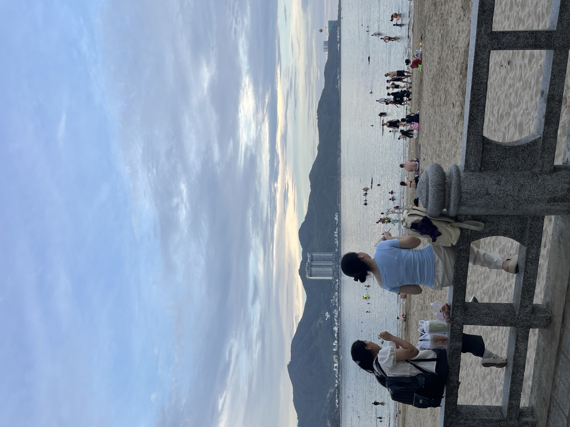 Two women sit on a stone wall looking out at the beach in Bac Nha Trang Ward, Vietnam. One woman wears a light blue shirt and white shorts, while the other wears a white shirt, dark pants, and a black backpack. They are both eating with chopsticks. Behind them is a beach populated with people swimming and relaxing on the sand. In the distance, mountains rise up along the coast. The sky is filled with clouds.