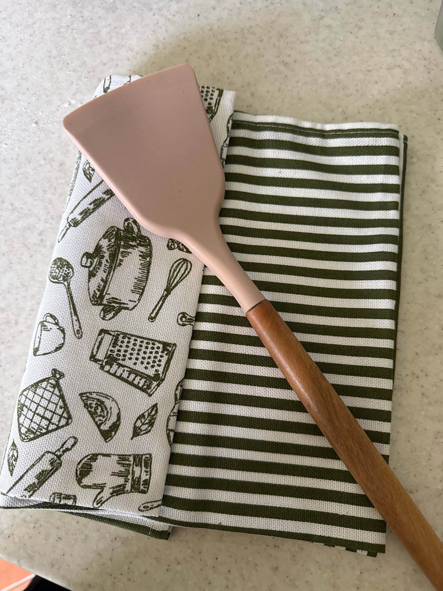 The image shows a kitchen spatula with a light pink silicone head and a wooden handle, resting on two folded kitchen towels. One towel has a white background with a dark green line pattern. The other towel is white with a dark green illustration of various kitchen utensils and food items. The scene is set on a speckled countertop, likely in a kitchen. There is no indication of the location being Kaliningrad, Russia, or any specific city. The image is a still life, with no people or actions depicted.