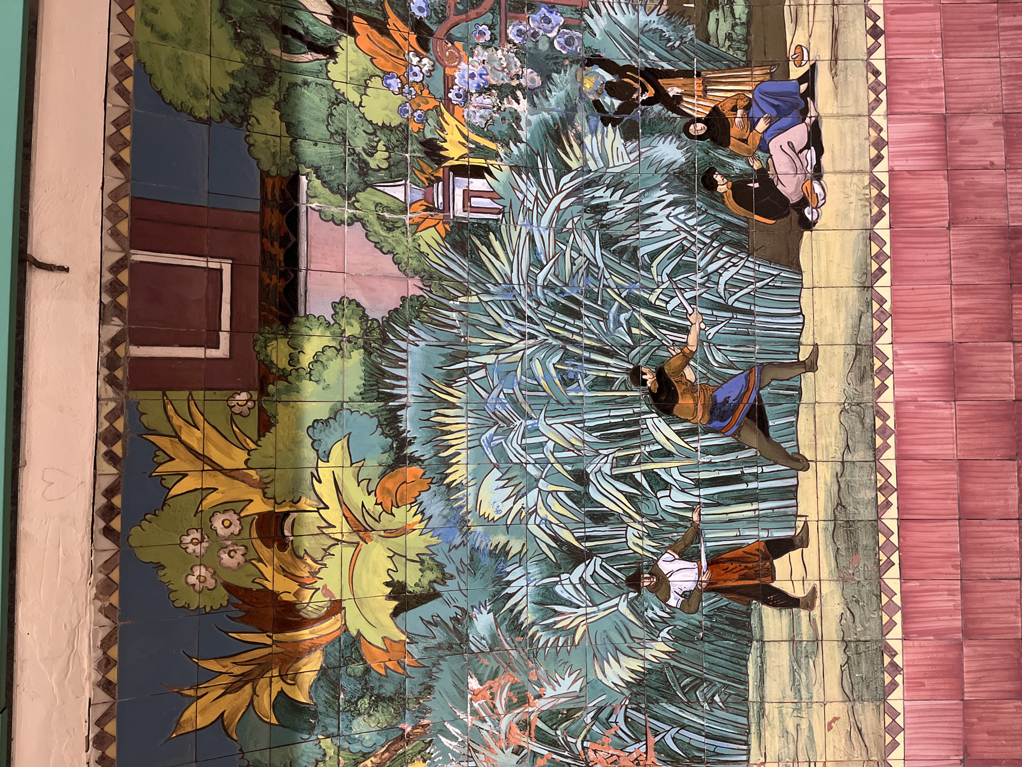 This media file displays a vibrant tile mosaic, likely an azulejo mural, depicting a dynamic scene within a lush natural landscape. The mosaic, when viewed horizontally, features at least five human figures in what appears to be traditional or historical attire.
On the right side of the mural, a man in a white shirt, brown hat, and reddish-brown garments, with dark hair, is shown in mid-motion, arms outstretched. Above him, a woman in a blue skirt and brown top, also with dark hair, dances or gestures with raised arms. Both figures are barefoot and appear to be engaged in a spirited activity.
Further to the left and slightly higher, a group of three figures is clustered. One woman, clad in a light blue top and pale pink lower garment, sits or crouches, facing away. Another individual, possibly male, in a black jacket and white shirt, interacts with her. A third figure, dressed in brown, observes or participates from behind.
The setting is characterized by abundant, stylized vegetation, including large green and yellow leaves, possibly palm fronds or bamboo-like plants, and smaller blue and white flowers. In the background, two small buildings are visible: a red-brown structure with a white-framed window on the far left, and a white, peaked-roof building above it. Patches of dark blue sky are seen between the foliage. The ground beneath the figures is a light brown/beige path, bordered on the far right by a reddish-brown tiled floor and on the far left by a decorative geometric border against a light wall.
The art style is illustrative and colorful, typical of ceramic murals, suggesting a daytime scene. The overall scene evokes a narrative or an event, possibly a celebration or a moment from a story, with an energetic and engaging atmosphere. There is no visible text in the image.