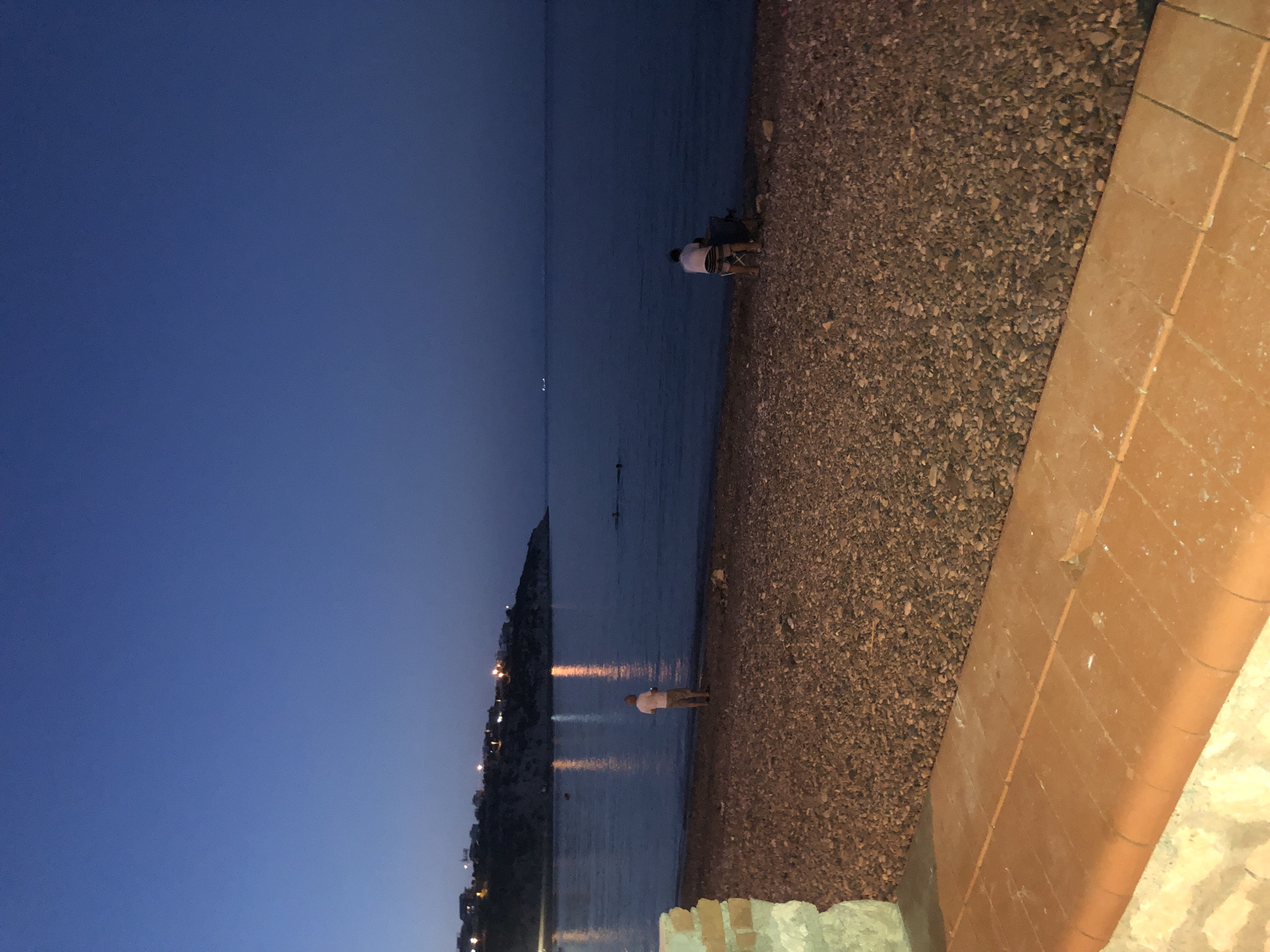 The image depicts a pebble beach at dusk in Karaburun, Turkey. The sky is a deep blue, transitioning to a lighter shade near the horizon. The sea is calm and dark, with reflections of lights from the shore visible on its surface.
On the beach, two individuals are present. One person, facing the sea, stands near the water's edge wearing a white shirt and shorts. Further down the beach, another person is seated in a folding chair, also facing the sea. In the background, houses with illuminated windows are visible on a hillside overlooking the bay. The foreground shows a textured orange tiled platform and a portion of a stone structure.