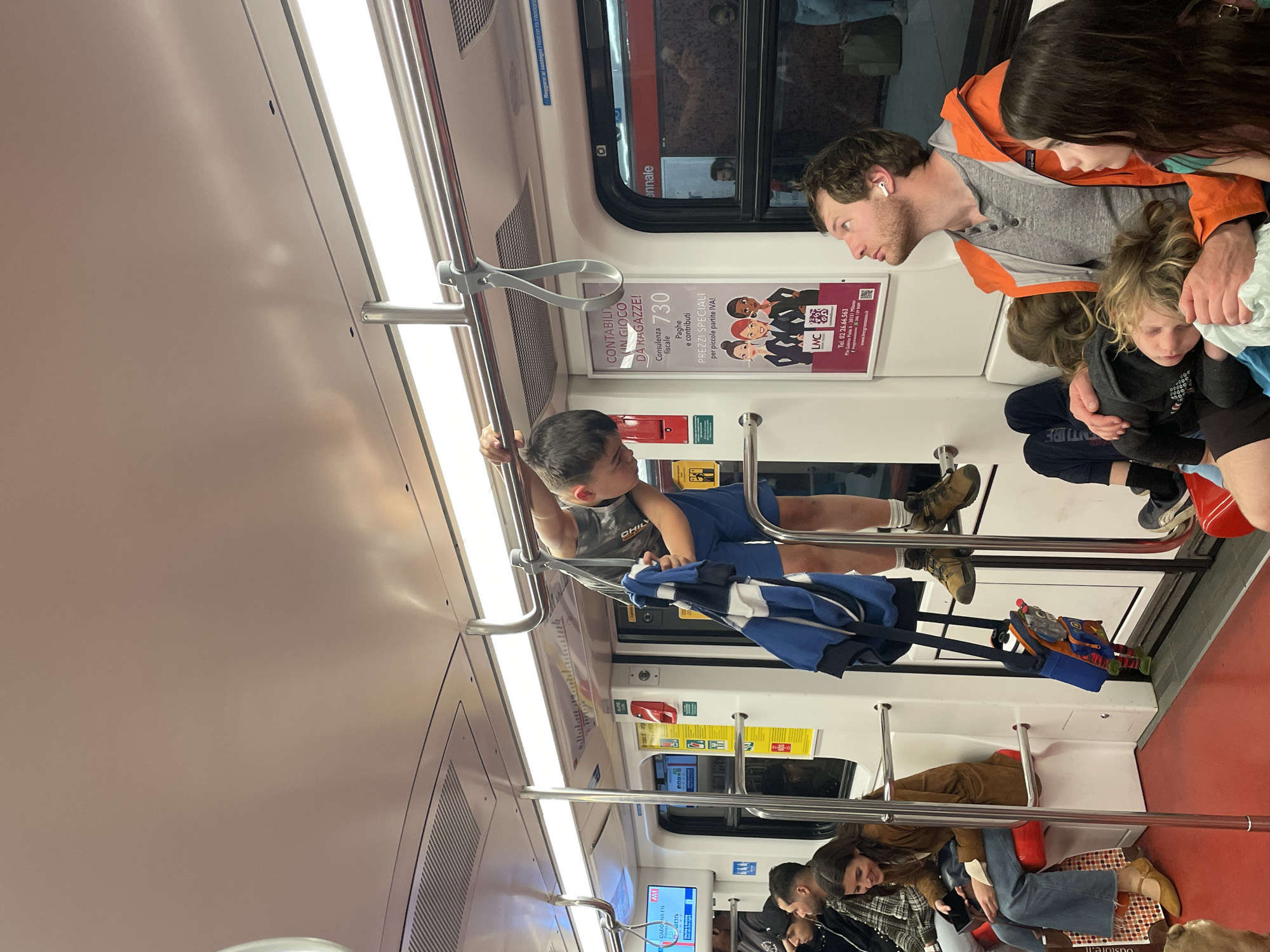 The photo captures a scene inside a Milan Metro train car. A boy wearing a blue shirt and shorts is grasping an overhead grab bar. An adult male with an orange jacket and an AirPod in his ear is standing nearby. Another adult woman and two children are also nearby. Several passengers are seated, including a woman in a brown jacket and jeans. The train's interior includes advertisements, emergency signs, and safety instructions. The advertisements include information about Contabili in Gioco, fiscal services and contact information, Ple Gabrio Pisla 6-20131 Milano.