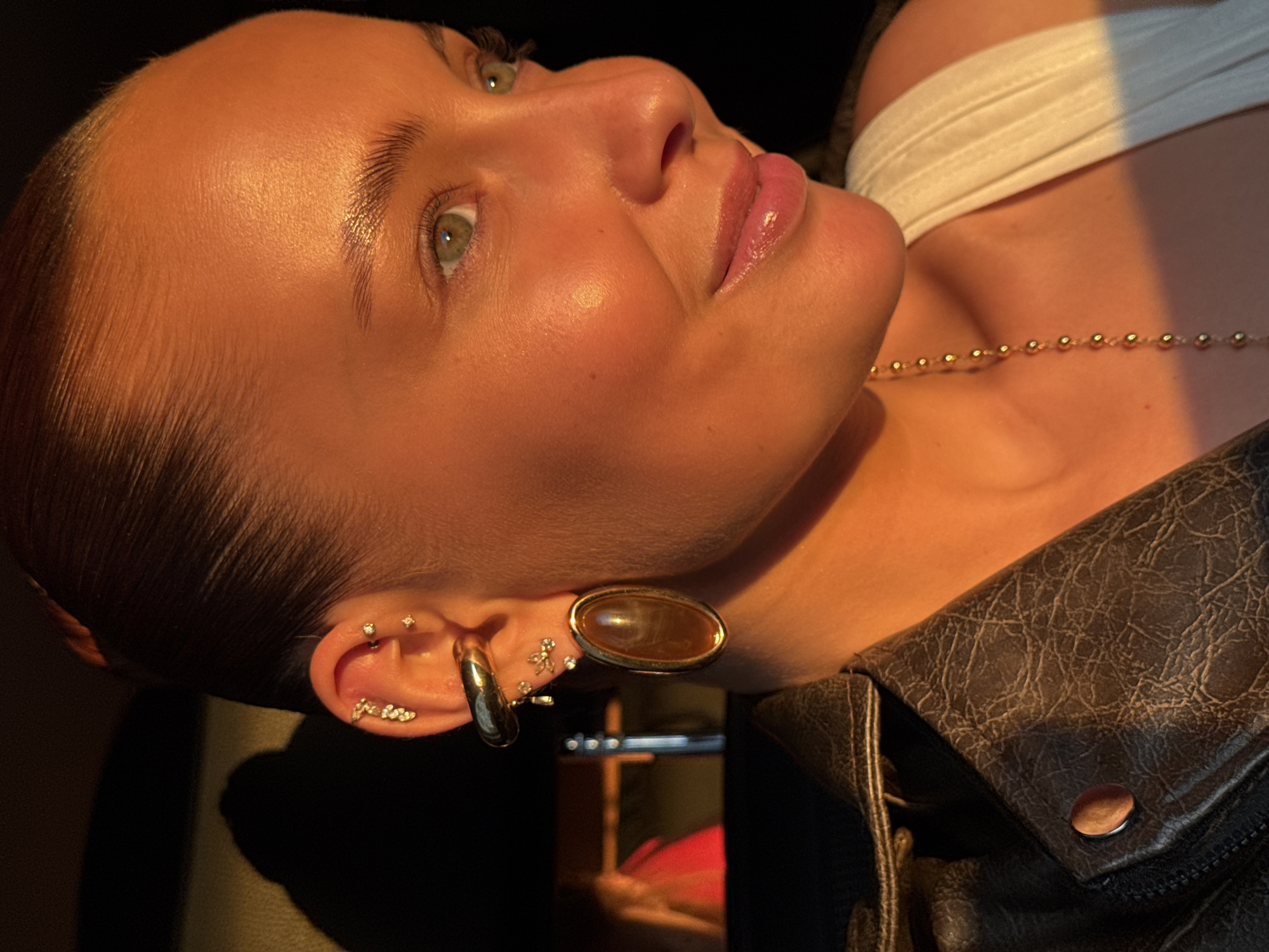 The media file is a close-up, side-profile portrait of a young woman, illuminated by warm, golden light, suggesting it was taken during the golden hour. Her skin appears dewy and has a healthy glow, with subtle makeup enhancing her features. Her green eyes are looking upwards and slightly to the right, and her lips are slightly parted, giving her a thoughtful or perhaps content expression.
She has multiple piercings on her ear, including small stud earrings with what appear to be diamonds, a hoop earring, and a larger, oval-shaped earring with a dark, amber-colored stone. She is wearing a delicate gold necklace with small beaded elements. Her hair is slicked back, accentuating her facial structure. She is also wearing a white garment, possibly a crop top or bandeau, and a dark, textured jacket, likely made of leather, which is partially visible in the foreground.
The lighting creates strong shadows and highlights, emphasizing the contours of her face and the texture of her clothing. The background is dark and indistinct, focusing all attention on the subject. There are no visible signs of the specific location in Chaikovsky, Russia, nor any indication of the weather. The overall mood is intimate and artistic, focusing on beauty and personal adornment.