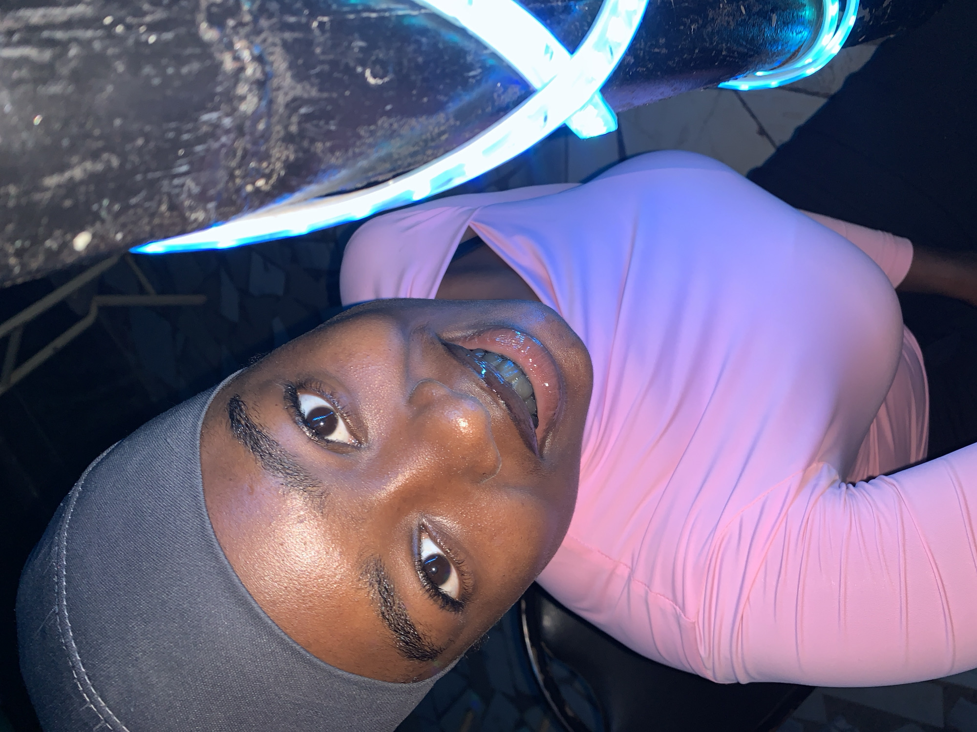 A close-up, tilted shot shows a dark-skinned person wearing a gray cap and a long-sleeved pink shirt or dress. The person looks up at the camera, their lips slightly parted and glossy. Above them, partially visible, is a surface with a dark, textured material and bright blue neon lights attached. The environment is dark, with glimpses of what appear to be tiled flooring in the background. The location is in the city of New Stadium, Nigeria. The scene is indoors, possibly at night, illuminated by the blue neon lights and the camera's flash.