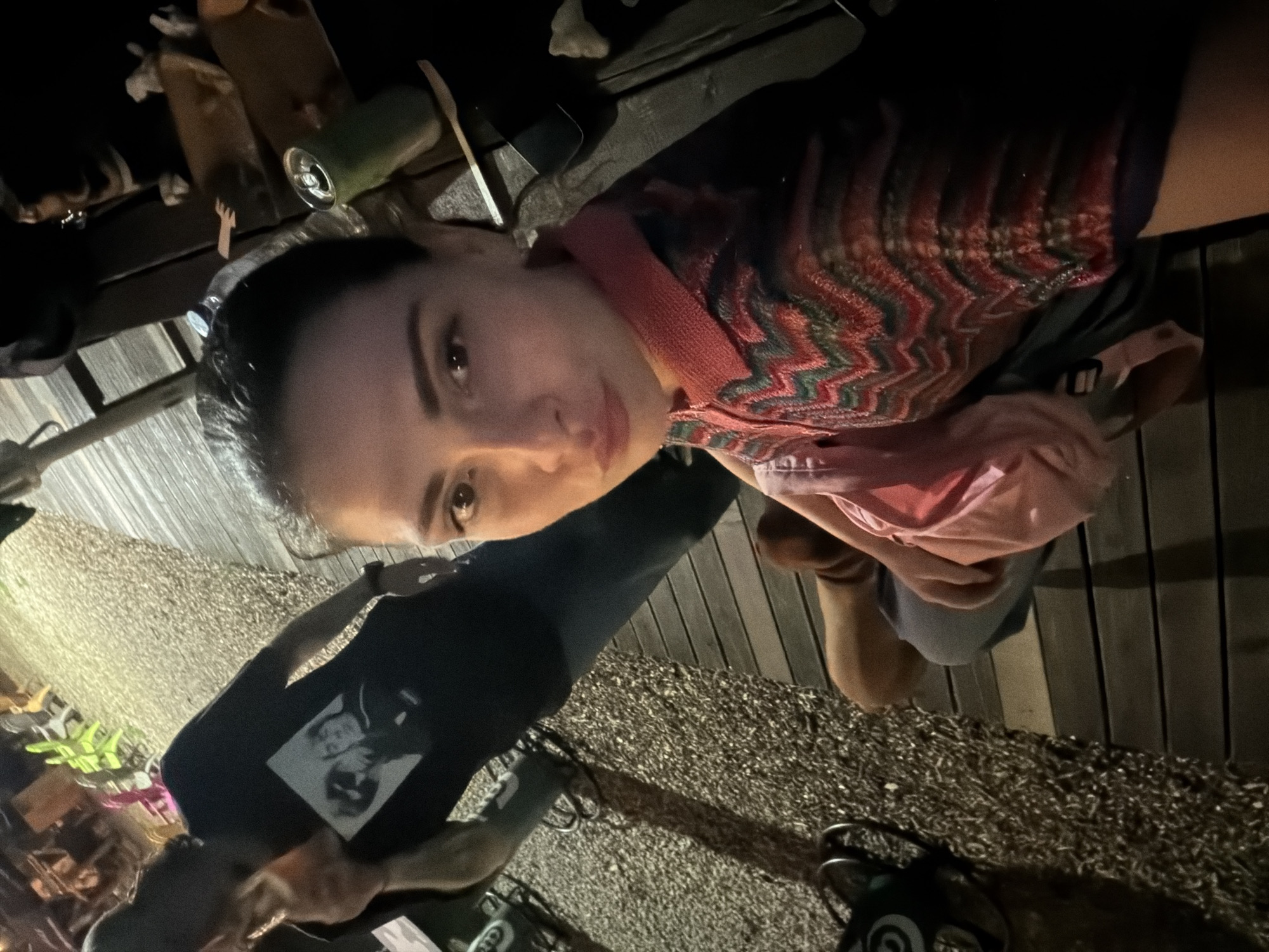 The image captures a low-light, outdoor evening scene, presented from a slightly tilted, low-angle perspective. The primary subject is a woman in the foreground, taking a selfie-style photograph. She has dark hair pulled back, with her left eye and the left side of her face visible. She is wearing a collared top with a vibrant zigzag or chevron pattern in shades of red, green, and possibly white, over a lighter, solid pink or salmon-colored shirt. Her lips are slightly pursed, and she looks directly at the camera. She is wearing dark blue or grey bottoms.
To her left, slightly behind her, a man stands, partially facing away from the camera. He has dark hair, possibly styled in dreadlocks or pulled back. He is wearing a black t-shirt featuring a prominent white graphic print of two figures, resembling an iconic music band like The Beatles. He also wears dark blue or grey trousers and a watch on his left wrist. He holds a clear glass or cup to his mouth, appearing to be drinking.
The environment suggests an urban outdoor social venue. The ground consists of two distinct surfaces: a light-colored, coarse gravel path occupies the left side of the frame, and a dark wooden plank deck or flooring covers the right side. On the wooden surface to the right of the woman, a green beverage can with an open pull-tab and a clear plastic water bottle with a blue cap are visible. In the far background, blurred figures of other patrons are seated at tables, suggesting a gathering. Artificial lighting illuminates the scene, creating shadows on the gravel and highlighting distant greenery. A dark metallic object, possibly part of a bicycle rack or outdoor furniture, is on the gravel to the far left. The ambiance and structural elements, including the combination of gravel and wooden decking, are characteristic of many relaxed outdoor evening establishments found in the city of Sofia, Bulgaria, such as popular garden bars, outdoor restaurant patios, or urban cultural spaces.