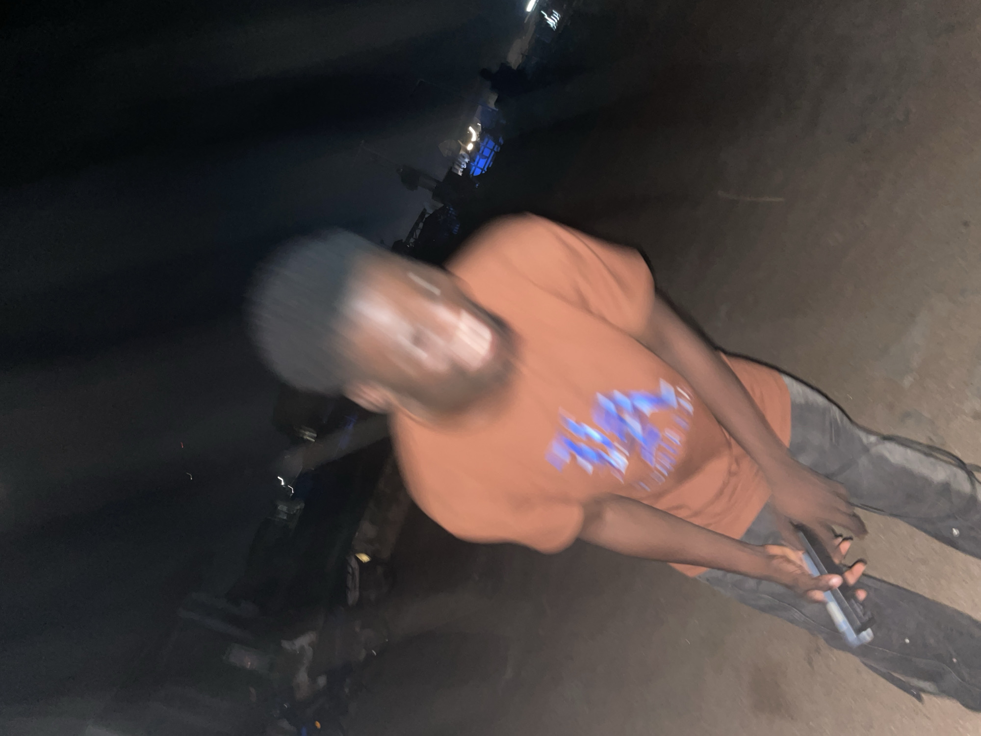 The image shows a young Black male, appearing to be in his late teens or early twenties, standing outdoors at night in what seems to be an unpaved, somewhat rough urban area. He is wearing an orange t-shirt with a blue graphic design on the front and dark pants. He is holding a silver mobile phone in his hands, positioned in front of him. The background is dark and blurry, with some distant lights and structures suggesting an outdoor setting in a built-up area, possibly a market or a street scene in Lokoja, Nigeria. The lighting is dim, and the image has a motion blur effect, making precise details of the surroundings indistinct. The overall impression is of an informal, possibly busy, nighttime environment.