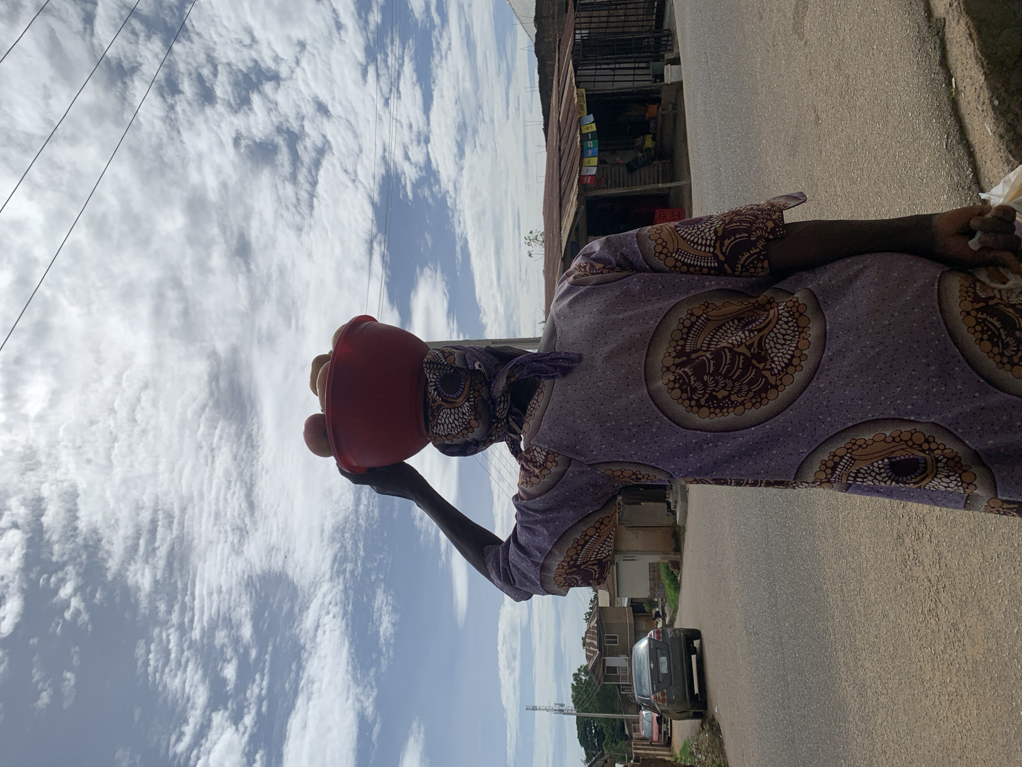 An adult individual, viewed from behind, walks on an asphalt road in Jos, Nigeria, under a partially cloudy sky. The person, with dark skin, wears a light purple garment adorned with large, patterned circular motifs in brown, orange, and white, and a matching headwrap. A red plastic bowl containing several light-colored, round fruits is balanced on the individual's head, supported by the raised right hand. The left hand holds a white plastic bag.
To the left of the road, a dark grey sedan is parked, facing away, with a partial view of a red vehicle behind it. Beyond the vehicles are residential structures, one light-colored with a brown roof, and green foliage leading to a distant communication tower. To the right, a small commercial structure with a rusted corrugated metal roof displays multiple colored items (likely merchandise) hanging from its facade. Utility wires traverse the upper right portion of the sky. The road surface appears worn.