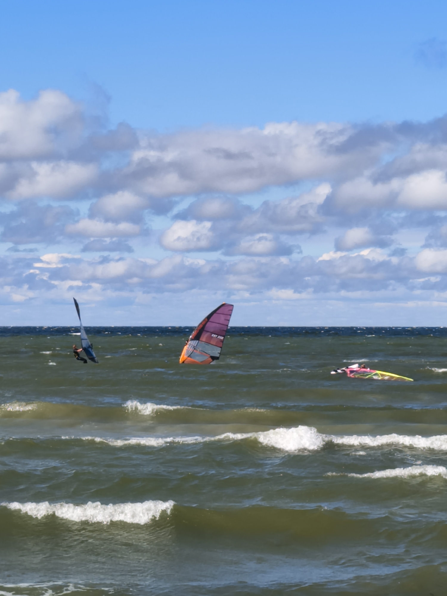 The image depicts a windy day at sea with three windsurfers on the choppy, dark green water. The sky is a bright blue with scattered white and gray clouds. In the foreground, waves with whitecaps break towards the viewer.
The windsurfers are positioned at different distances from the camera. The closest windsurfer, to the left of the frame, is actively engaged in the sport, with their sail raised. Another windsurfer, slightly further to the right and more central, is also on their board with a colorful sail. The third windsurfer is furthest to the right and appears to be in the process of turning, with their colorful sail extended.
This scene is consistent with activities that might take place at a popular windsurfing spot in Tallinn, Estonia, such as Pirita Beach or other coastal areas known for water sports, especially during favorable weather conditions. The open sea, choppy waves, and clear sky suggest an outdoor recreational setting.