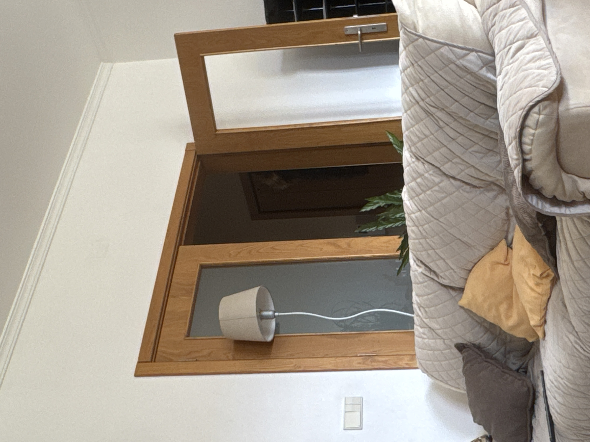 The image shows a wooden window with an open top section, mounted on a white wall. Inside the window frame, there is a cylindrical object with a cord extending from it. Part of a heavily quilted, light-colored sofa or bed is visible to the right of the window, with a brown pillow and a yellow cloth item resting on it. Below the window, there is a light switch on the wall. The location, Mirandela, Portugal, cannot be verified from this image.