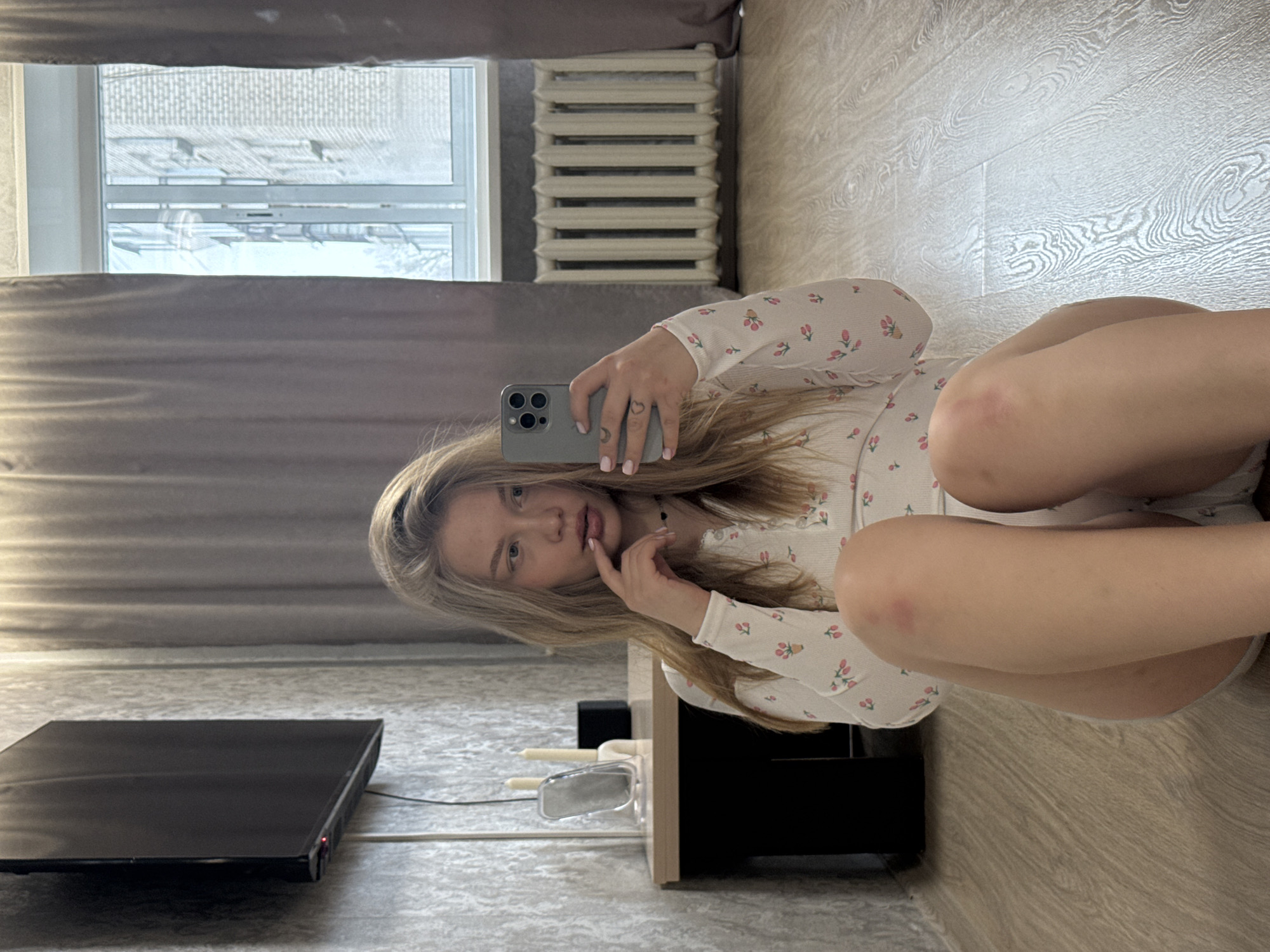 A young woman is taking a selfie in a room. She is kneeling on a wooden floor with her legs tucked to the side and is holding a phone in her right hand, pointed at her reflection. The woman has light skin and blonde hair. She is wearing a long-sleeved white shirt with a red floral pattern. Behind her, there is a wall with a TV and a small cabinet. To the left, there is a window with curtains partially drawn. Through the window, a building is visible, suggesting an urban environment. The location is Chaikovsky, Russia.