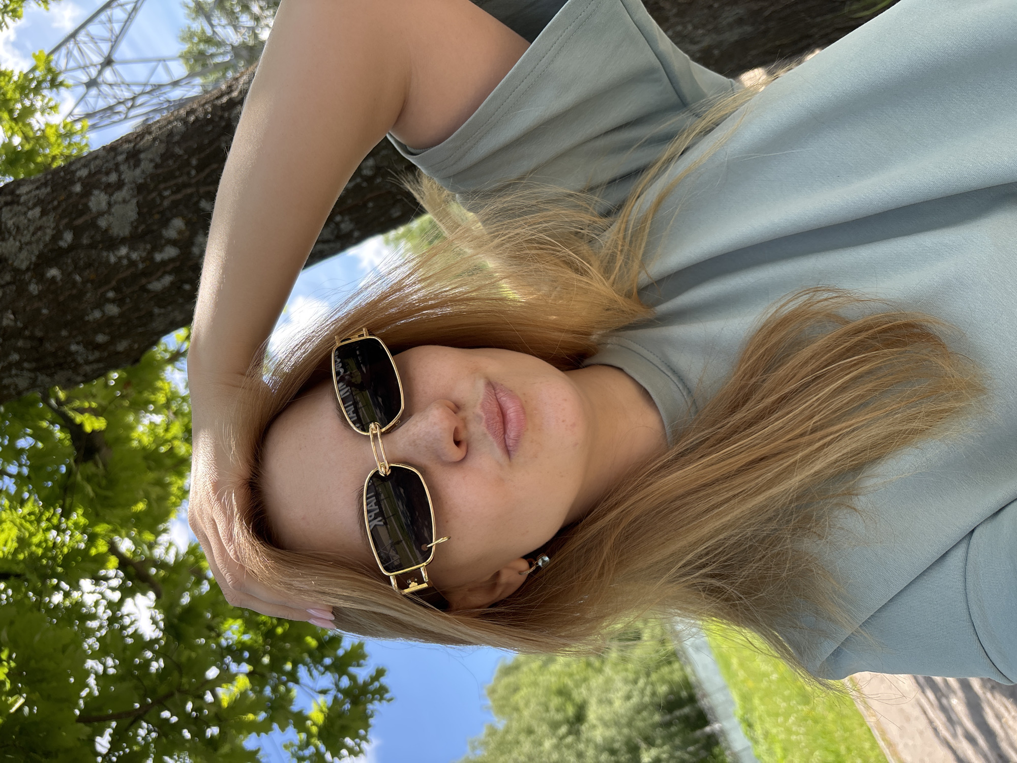 This close-up, rotated selfie features a person with light-colored (blonde or light brown) hair, sunglasses, and a light blue/mint green t-shirt, set against a vibrant outdoor backdrop. The person's head is tilted back, and one arm is raised, with their hand resting near their hair.
The subject is wearing gold-framed, rectangular dark sunglasses, with the word "XYRAL" (or similar, reversed) faintly visible on the inner surface of one lens. A small, dark stud earring is visible in their right ear. Their lips are slightly parted, suggesting a relaxed expression, though the sunglasses obscure their eyes.
The setting is outdoors, under a bright blue sky with scattered wispy clouds. Lush green trees and foliage are prominent in the background, hinting at a park or natural area. Through the foliage in the upper left, a large, intricate metal lattice structure, possibly part of a tower or bridge, can be seen. The bright sunlight suggests it is daytime, likely late morning or afternoon, with clear weather. The ground below the trees, visible in some crops, appears to be green grass and a paved path.
The overall impression is a casual, sunny day portrait. The location context provided is Rusanovo, Russia.