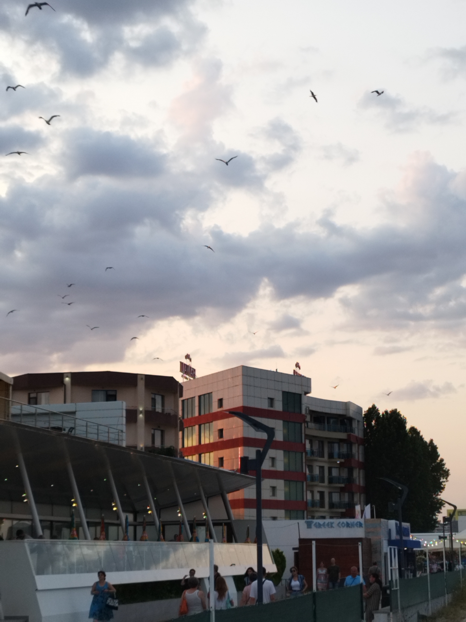 This media file depicts a scene in Constanța, Romania, likely during the late afternoon or early evening, as suggested by the warm, soft lighting and the pastel hues in the sky. The weather appears to be partly cloudy, with dramatic, dark clouds interspersed with lighter, more translucent ones, creating a dynamic sky. A flock of birds is scattered across the sky, adding a sense of movement and life to the scene.
The foreground features a building with a modern, open-air structure supported by numerous angled beams, possibly a restaurant or a public gathering space with outdoor seating. People are visible walking and standing in front of and within this structure, suggesting a relaxed, public atmosphere. Some individuals are captured in mid-stride, while others appear to be socializing.
Behind this structure, several multi-story buildings rise, with varying architectural styles. One building prominently displays red and white horizontal bands, contrasting with its glass windows. Another building has a more muted, beige facade. A sign with the text "GREEK CORNER" is visible on one of the lower buildings, indicating a potential establishment or restaurant name. In the background, a dense cluster of trees adds a touch of nature to the urban landscape. The overall mood conveyed by the image is one of tranquil urban life set against a striking sky.