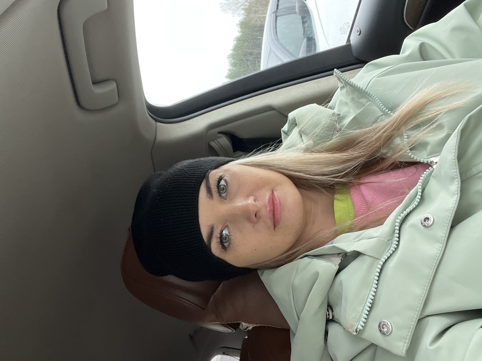 A woman with light complexion, wearing a black beanie, a pastel green winter jacket, and a pink top with a green stripe, is lying down in the back seat of a car. The interior of the car includes a brown leather headrest, a grey ceiling, and a grab handle. Through the car window, there is a partial view of another vehicle and trees. The image was taken in the city of Chaikovsky, Russia.