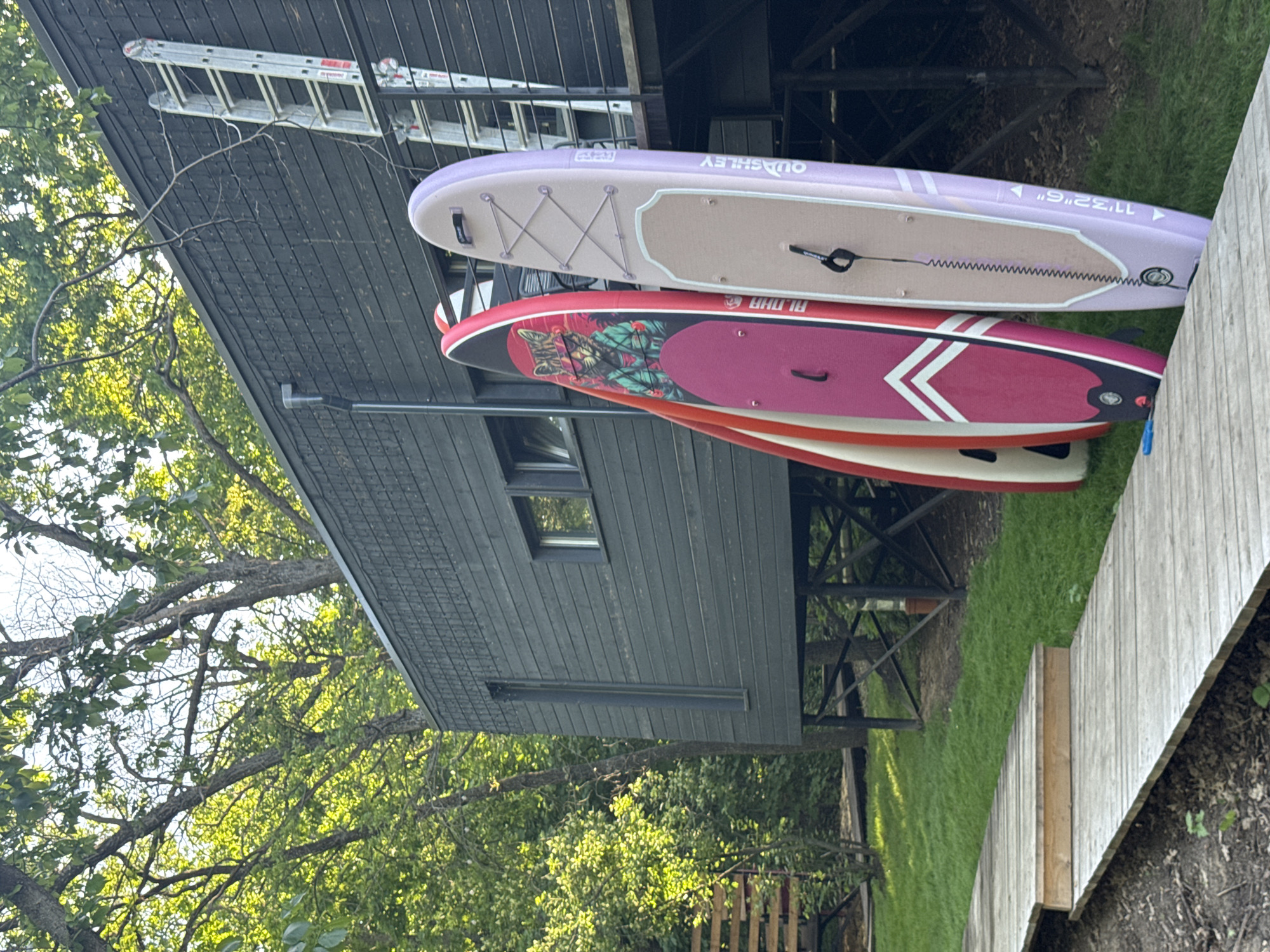 The image shows a dark gray, elevated house or building on stilts, likely near water, with several paddleboards leaning against it. A silver ladder rests on the roof of the building. The paddleboards are various colors and designs, including pink, red, and purple. The building is situated near a wooden boardwalk. The location is not confirmed to be Усть-Курдюм, Russia, as the image does not provide any indicators for location. The environment includes greenery and trees. There are no people visible in the image.
