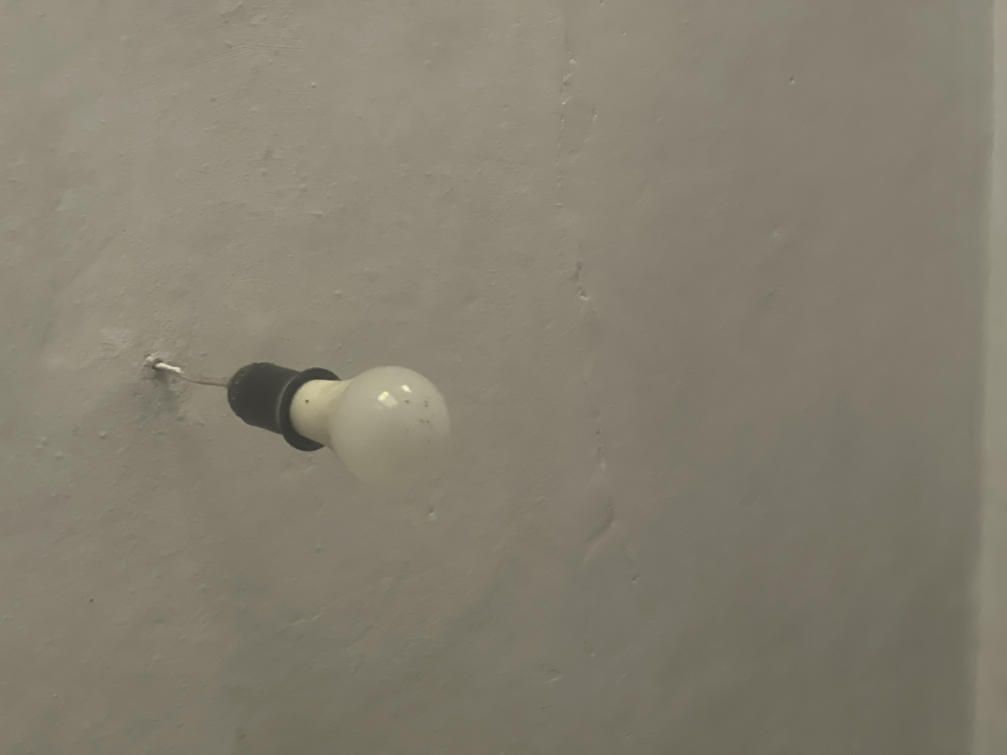 The image shows a bare light bulb hanging from a cord attached to a textured, light-colored ceiling. The light bulb has a black socket and a white, round bulb. The ceiling appears to be made of concrete or plaster and has some imperfections. The overall lighting is dim and the scene is static. The location of Ohuivka, Ukraine is not depicted in the image.