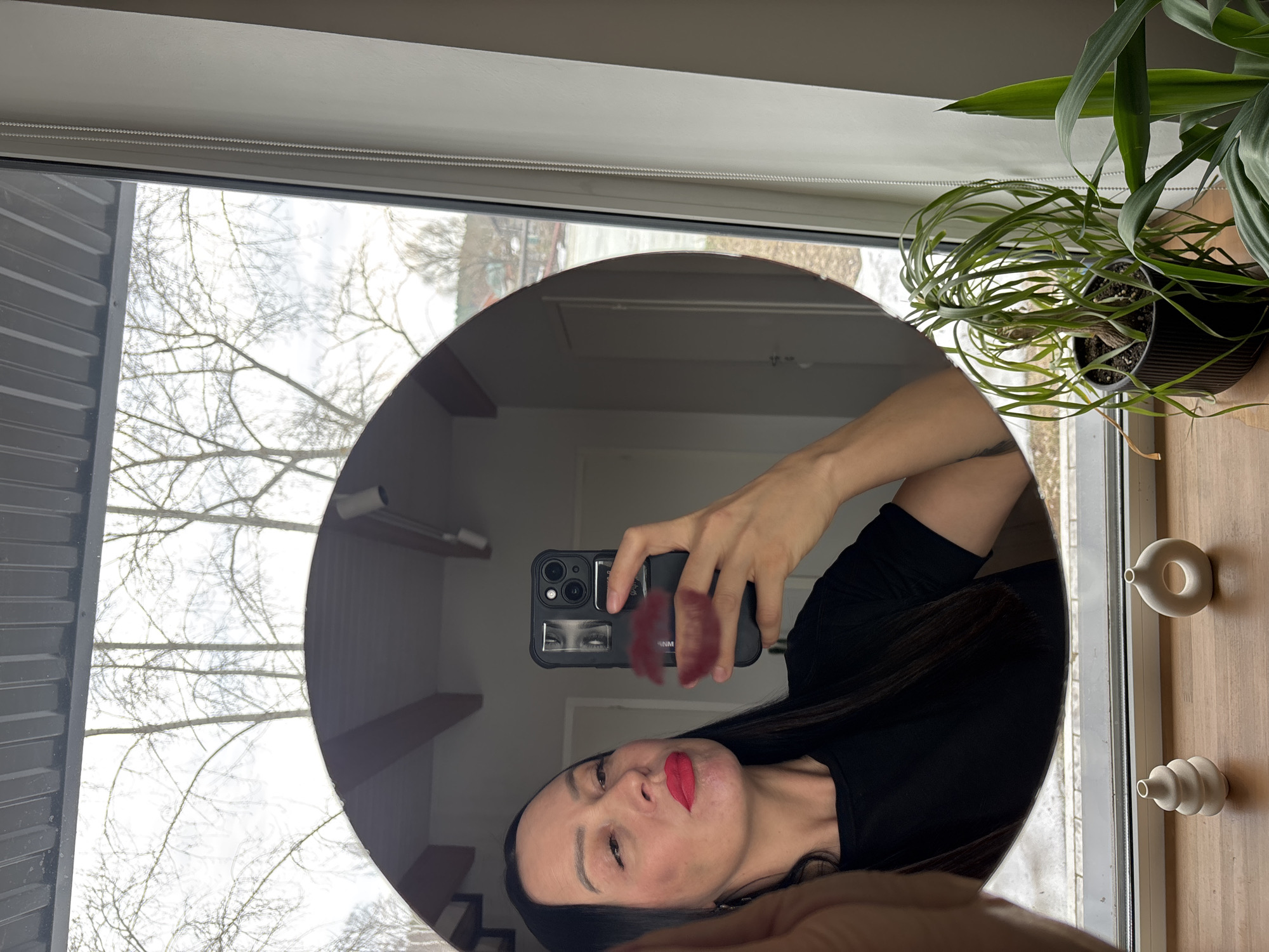 A woman is taking a selfie in a round mirror. The mirror is positioned in front of a window through which bare tree branches can be seen. A potted plant and two small, stylized vases are on a surface to the right of the mirror. The woman is wearing a black shirt and has dark hair. Her lips are painted red, and she is holding a black smartphone with a lip-shaped sticker on the back. The location is in an apartment, likely in the city of Chaikovsky, Russia based on the window frame and architectural details visible in the reflection.