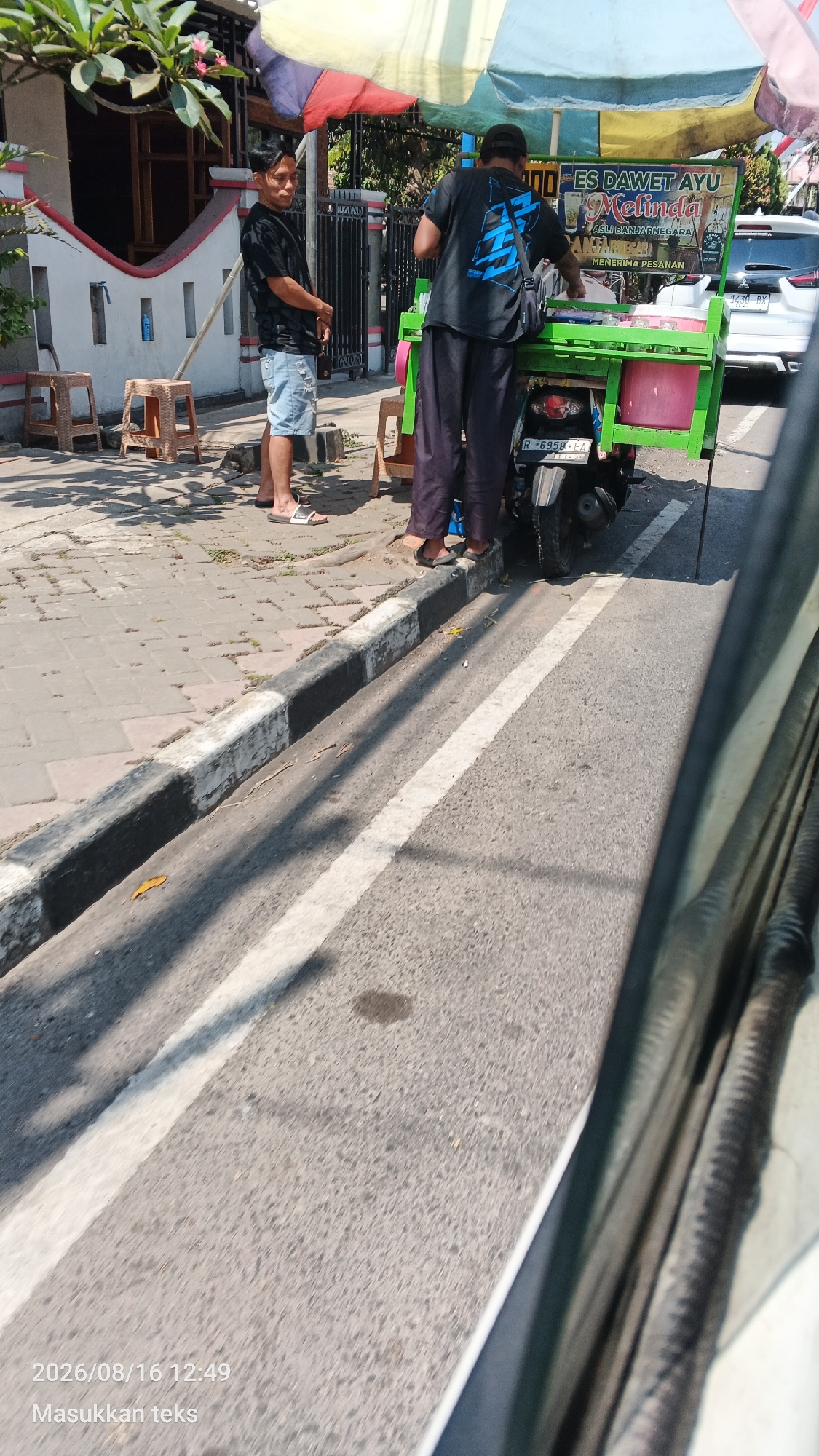 A man in a black t-shirt and shorts stands by a street food cart on a sunny day in Pakis, Indonesia. The cart, which is green, is mounted on a motorcycle and sells "Es Dawet Ayu Melinda." Another man, wearing a black t-shirt with a blue graphic and dark pants, is attending to the cart, serving a customer. Two small wooden stools are placed on the sidewalk near the first man. The street has a white line marking and a curb with black and white paint. A white car is visible in the background, and a building with a white wall and red trim is to the left.