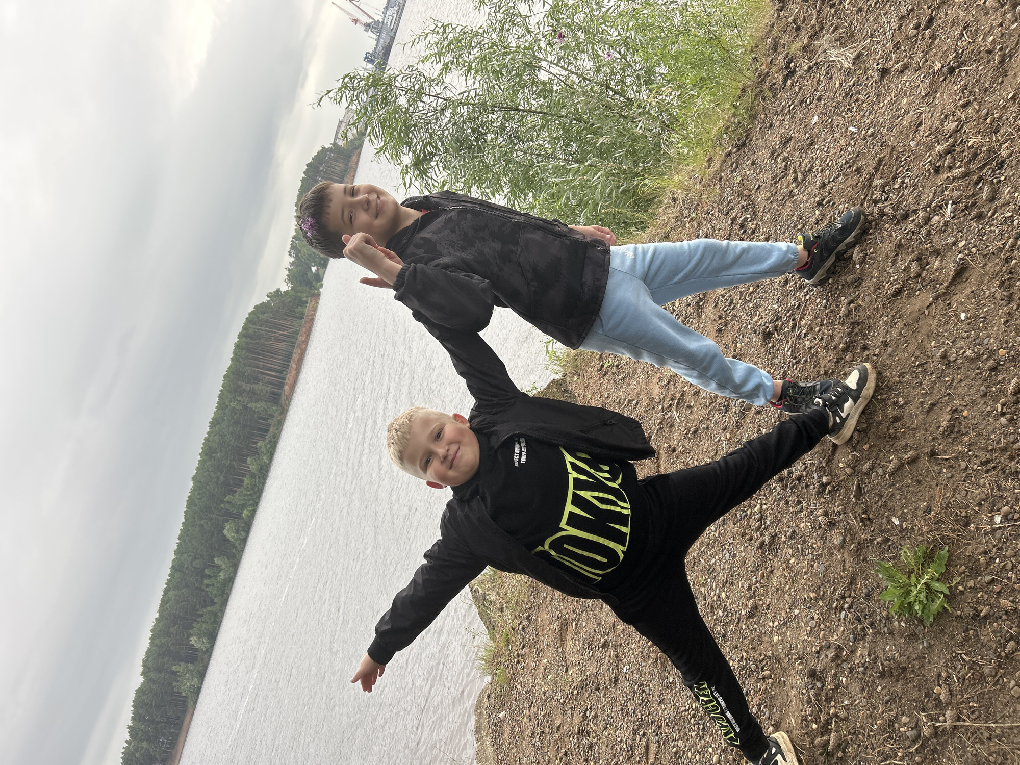 Two children are posing for a photo outdoors. The child in the foreground is a young boy with blond hair, wearing a black jacket and black pants with neon yellow lettering. He is smiling and has his arms and legs spread wide. The child in the background is a slightly older girl with dark hair, wearing a black patterned jacket and light blue pants. She is also smiling and has a flower crown on her head.
They are standing on a dirt and gravel embankment overlooking a body of water. In the distance, across the water, is a dense forest of pine trees. Further in the background, industrial cranes are visible, suggesting a port or industrial area. The sky is overcast and appears to be a cloudy day. The overall mood seems to be playful and happy. The visible text on the boy's pants reads "QUEEN" in large neon yellow letters.