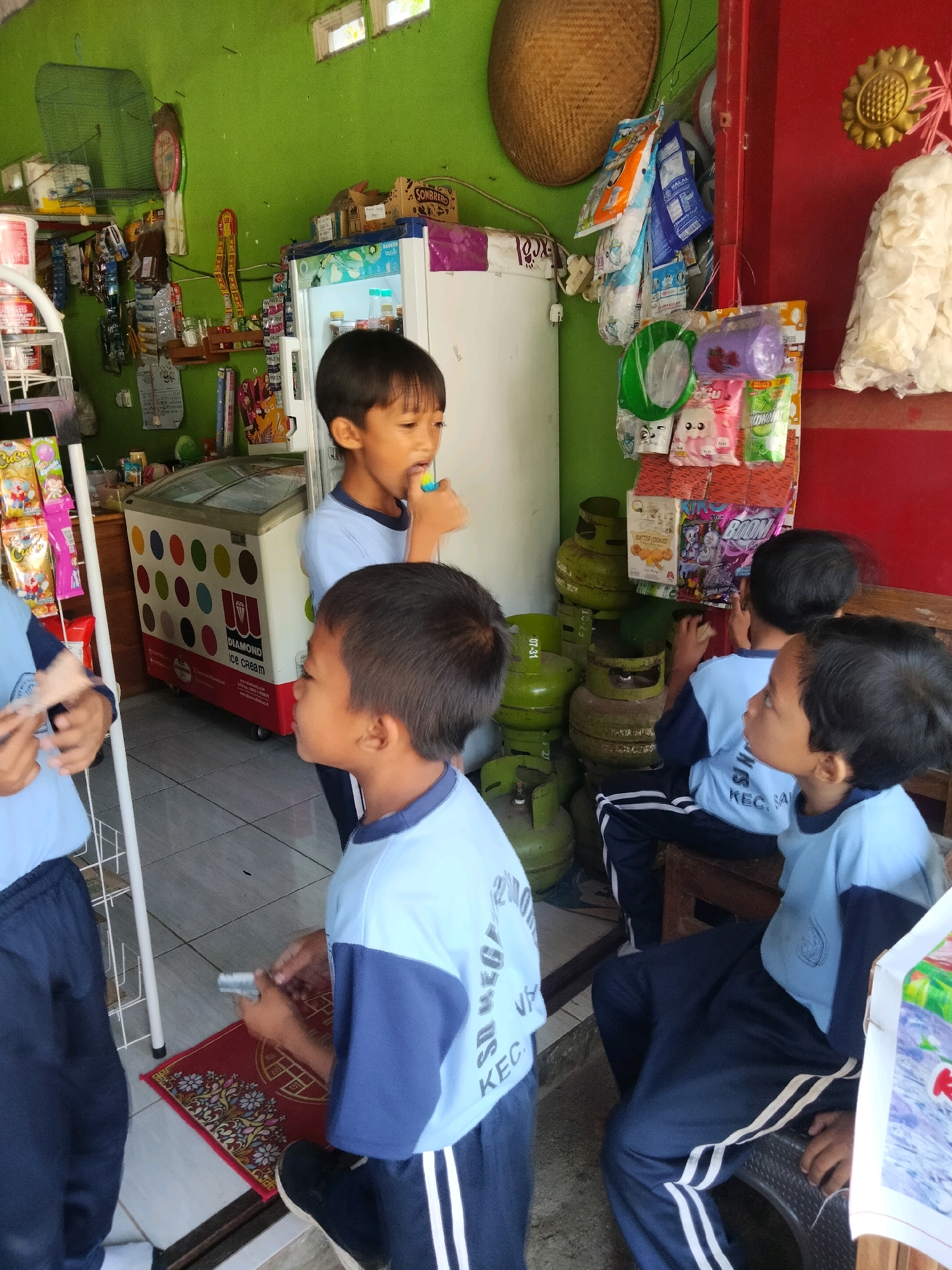 In the city of Sidorejo, Indonesia, at a small, open-air shop, a group of children in light blue and dark blue school uniforms are gathered. The shop is stocked with various goods, including bottled drinks, snacks, and household items. A white and colorful refrigerator with the "Diamond Ice Cream" logo is visible, suggesting it sells frozen treats. Green gas cylinders are stacked near the counter, and bags of chips and other packaged goods are displayed on shelves. One child is holding what appears to be a lollipop or a similar confection. Another child is examining a small rectangular object, possibly money or a ticket, in their hands. The scene is lively and informal, with the children seemingly engaged in shopping or making purchases.