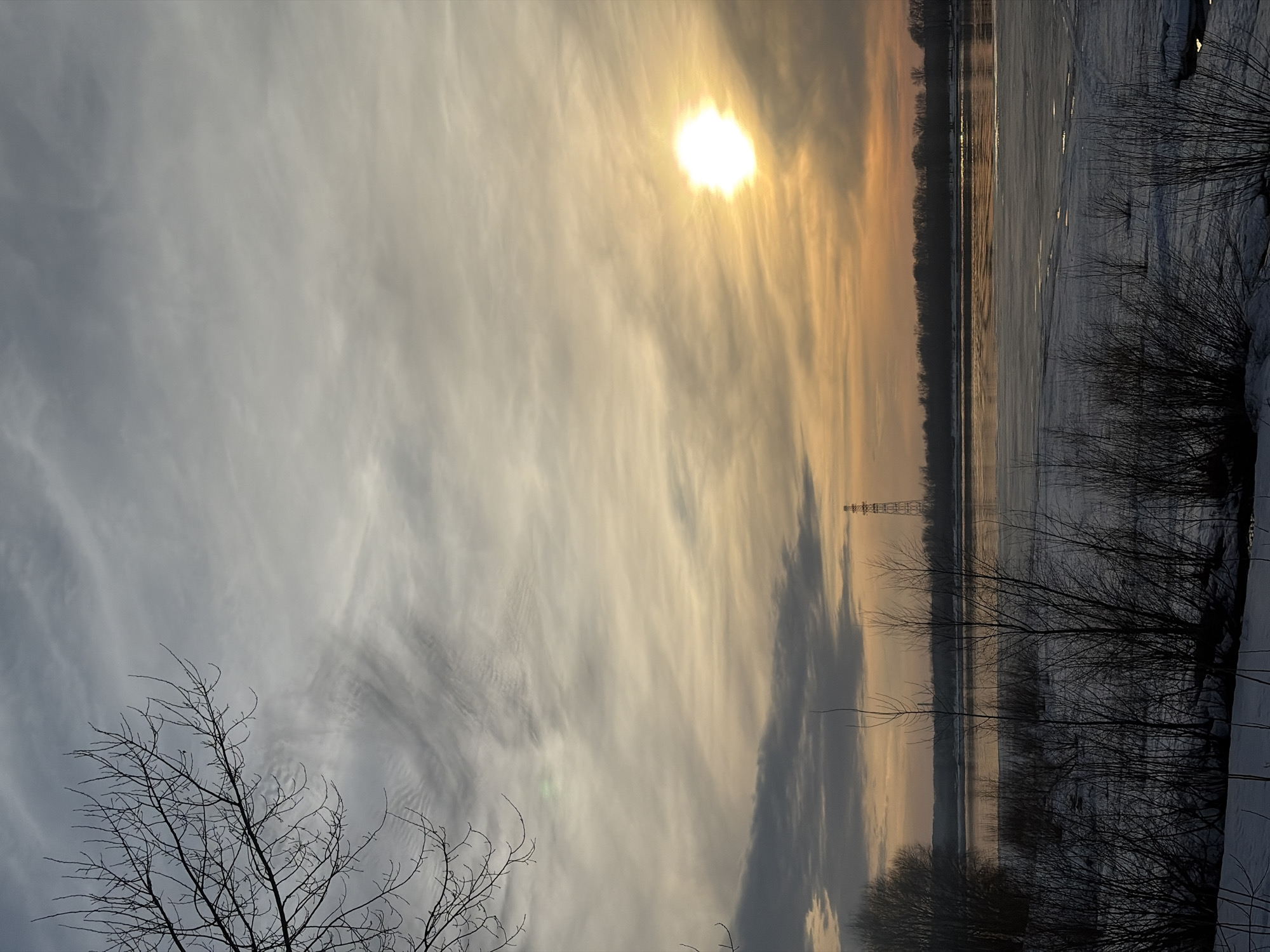 The image depicts a static landscape scene in Chaikovsky, Russia, during what appears to be either sunrise or sunset in late winter or early spring. The foreground consists of snow-covered ground with patches of dark earth and numerous silhouetted leafless bushes, their thin branches extending upwards. In the midground, a wide body of water, likely the Kama River, is partially covered by sheets of ice and slush, particularly closer to the viewer. Open water reflects the warm orange and gray tones of the sky. The background features a dark, dense line of bare trees along the opposite riverbank, stretching horizontally across the frame. A tall, lattice-style transmission or communication tower stands silhouetted against the horizon line on the far bank, positioned slightly right of center. Further to the left on the distant bank, faint industrial structures are visible. The sky, occupying the upper two-thirds of the image, displays a blend of gray, white, and orange cirrus and stratus clouds. The sun, a brilliant white orb, is prominently positioned in the upper right quadrant, casting an intense glow and warm hues across the sky. Bare tree branches from an unseen tree frame the upper left corner of the image. No individuals or specific actions are observed.