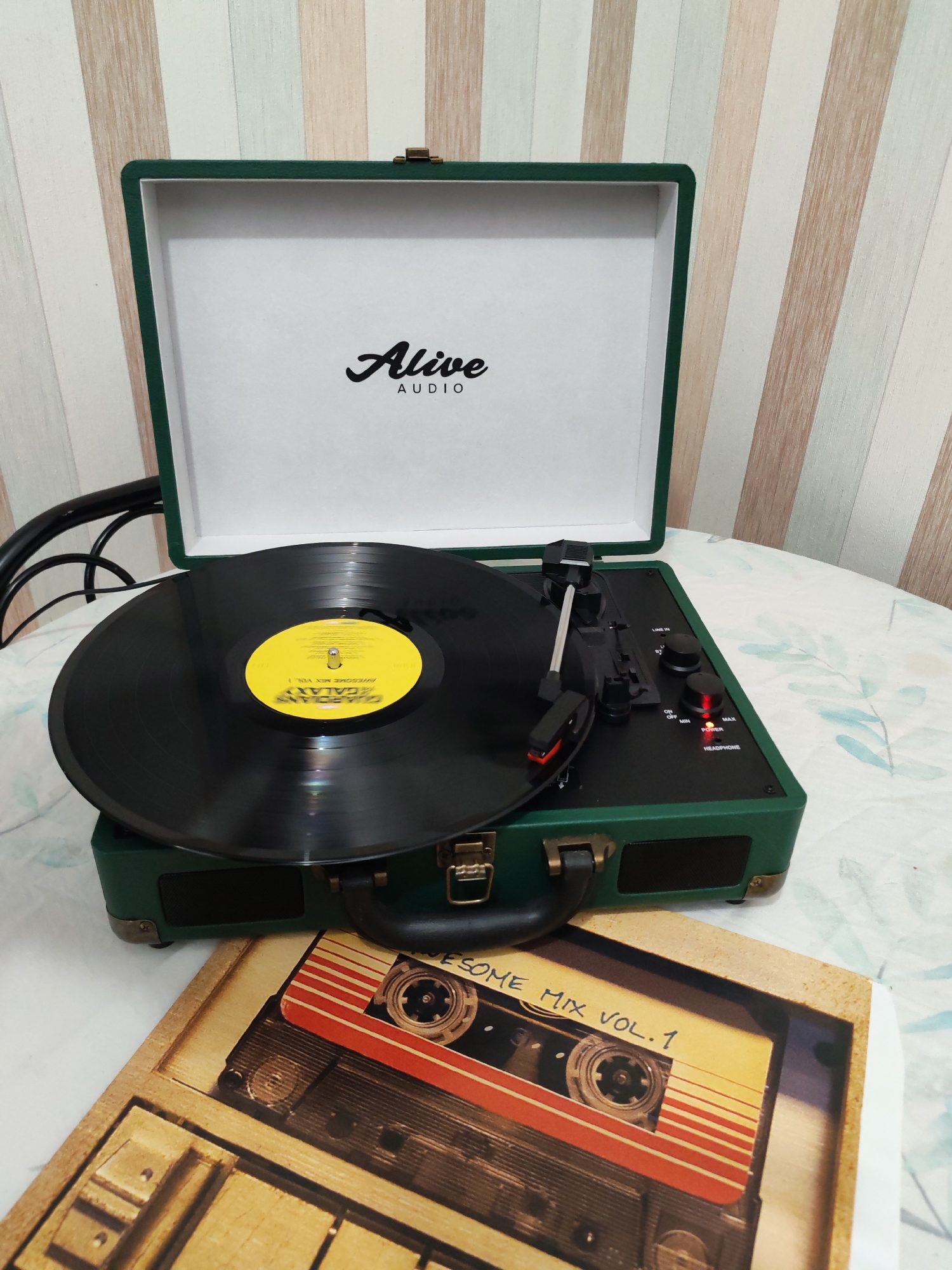The image displays an open, dark green portable record player, branded "Alive AUDIO," set up in an indoor environment, possibly a home in Izhevsk, Russia. A black vinyl record with a yellow label is positioned on the turntable's platter, featuring visible text "GUARDIANS OF THE GALAXY" and "AWESOME MIX VOL. 1." The record player's lid stands upright, revealing the brand logo, while its control panel on the right side features black knobs for "VOLUME" and "SPEED," an "ON OFF" switch, a red illuminated "POWER" light, and a "HEADPHONE" jack. The tonearm is resting over the record, indicating readiness for playback.
In the foreground, resting on the same surface as the record player, is a flat item, likely a book cover or a large coaster, designed to resemble a retro cassette tape. This item also prominently displays the text "AWESOME MIX VOL. 1," matching the record's theme and tying into the "Guardians of the Galaxy" franchise. The cassette design features iconic red and yellow stripes.
The record player and the cassette-themed item are placed on a white tablecloth adorned with a subtle, light blue and green leaf pattern. The background consists of a wall covered in vertical striped wallpaper in muted tones of beige, light brown, light green, and white. A black power cord is partially visible extending from the left side of the record player. The overall scene suggests a relaxed atmosphere of enjoying music through classic audio formats.