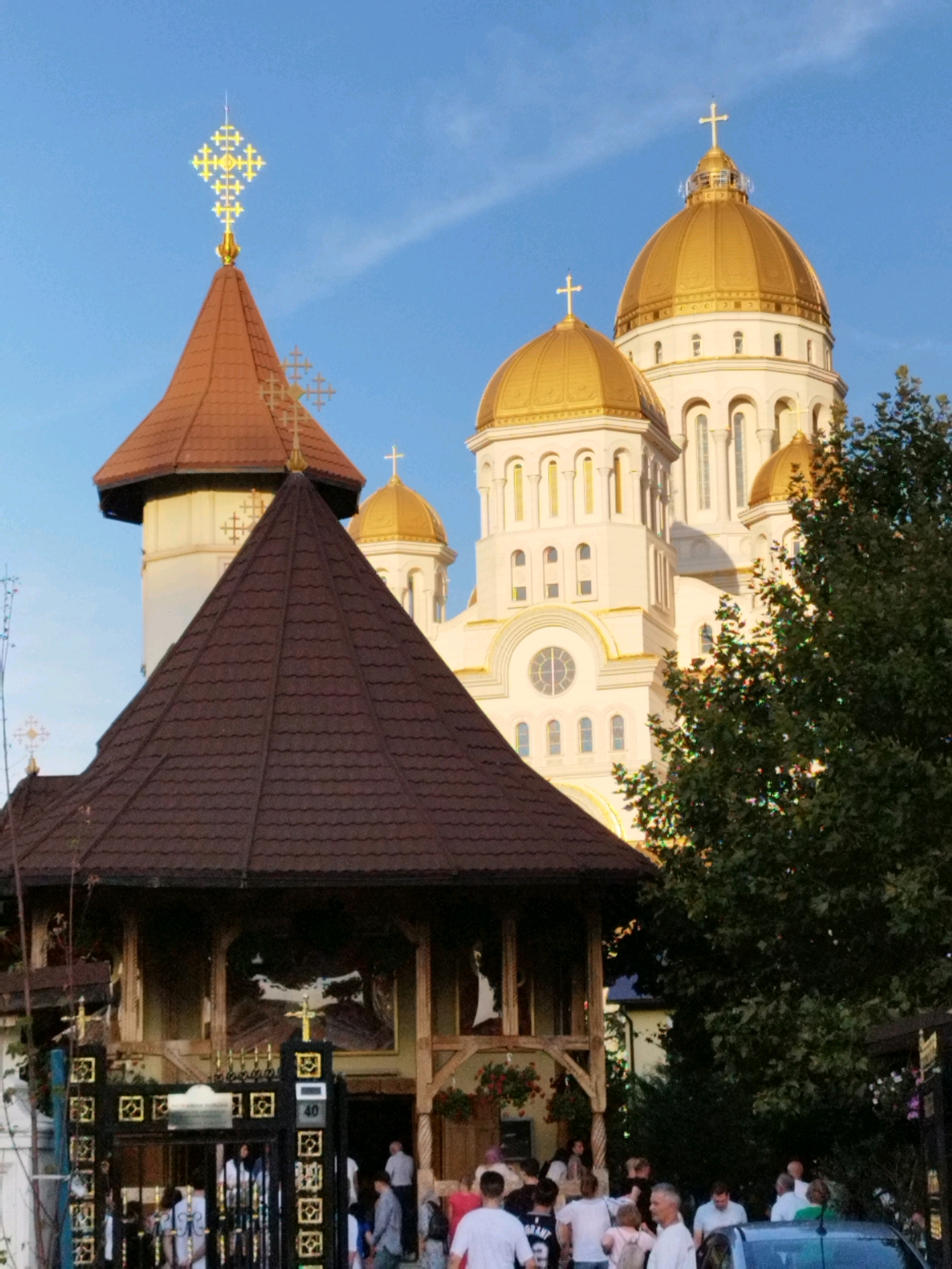 The image shows a view of the Romanian Orthodox Metropolitan Cathedral in Bucharest, Romania, on a clear day. In the foreground, a smaller brown-roofed building with an entrance gate is visible, with a crowd of people gathered. Behind this, the large white cathedral with golden domes and crosses is the dominant structure. Green trees are also present on the right side of the image. The sky is blue with some wispy clouds.