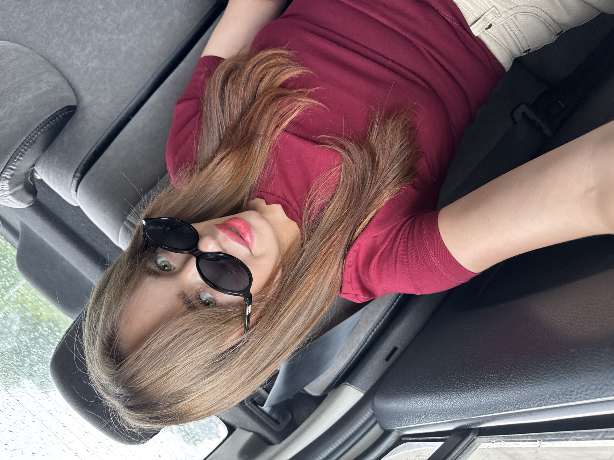 A woman with long, reddish-brown hair and green eyes is sitting in the back seat of a car. She is wearing black sunglasses, a dark red t-shirt, and light-colored pants. The car's interior is visible, including the seatbelts and car seats. The window shows a rainy outdoor scene, suggesting the car is in Russia, possibly Chaikovsky, Russia, given the context.